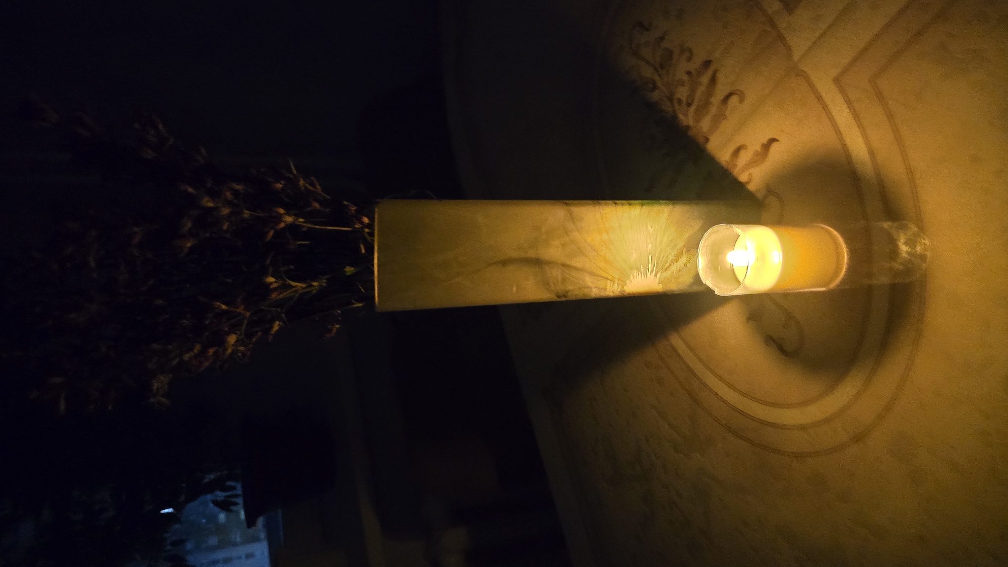 A lit candle sits on a table in a dimly lit room, casting a warm glow. The candle is in a clear glass holder and has a yellow flame. Behind the candle, a tall, rectangular vase filled with dried plant material is illuminated by the candlelight, creating a shadow on the table. The table surface has a textured pattern. The background is dark and out of focus, suggesting an indoor setting at night. The scene is located in Chaikovsky, Russia.
