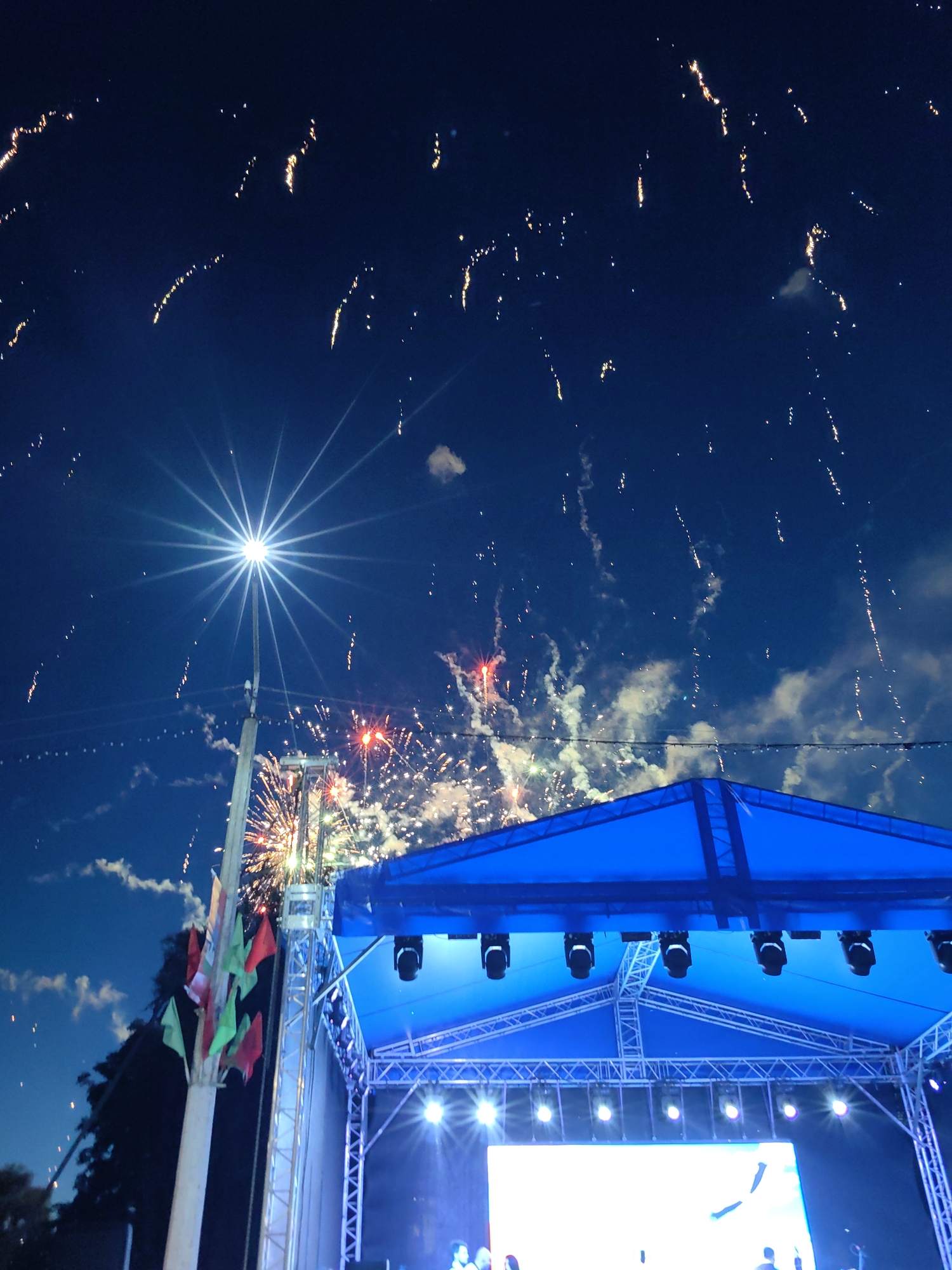An outdoor fireworks display is underway above a blue-roofed stage. The sky is a deep blue, with numerous streaks and bursts of light from the fireworks. A bright, star-shaped light source emanates from a pole on the left side of the image, likely a street lamp. Flags in red and green are attached to the same pole. The stage has a metal truss structure supporting a large blue tarp and a screen displaying a bright image. Several spotlights are visible on the stage structure. The overall scene depicts a festive nighttime event. The image was taken in the city of Myadzyel, Belarus.
