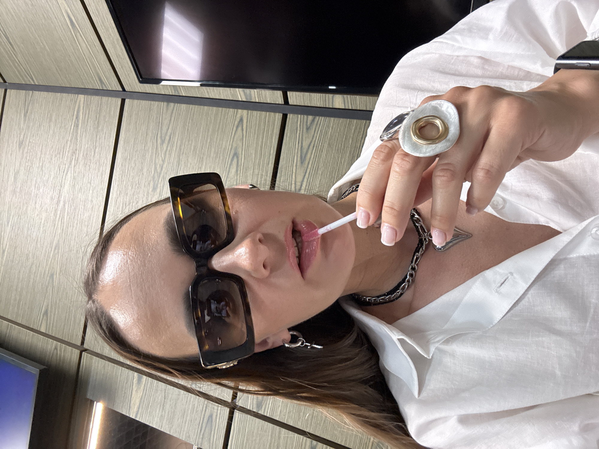 This is a vertical, close-up, medium shot of a person applying lip gloss. The subject is wearing large, square, tortoiseshell-framed sunglasses. Their lips are slightly parted as they hold a lip gloss wand with their right hand, which also wears a large, abstract silver and gold ring. A thick, silver chain necklace with a heart-shaped pendant is visible around their neck. They are wearing a white collared shirt, and the collar is partially unbuttoned. A black smartwatch is on their left wrist.
The background consists of light-colored wooden paneling. A dark, flat-screen television is visible at the top of the frame, and a sliver of a blue screen is in the bottom left corner. The lighting suggests it is daytime, possibly indoors with ambient light. The overall mood is casual and focused on personal grooming. The presence of a television and what appears to be office-like wood paneling suggest an indoor setting, possibly a home or office. There is no visible text in the image. The location context provided indicates Sochi, Russia.