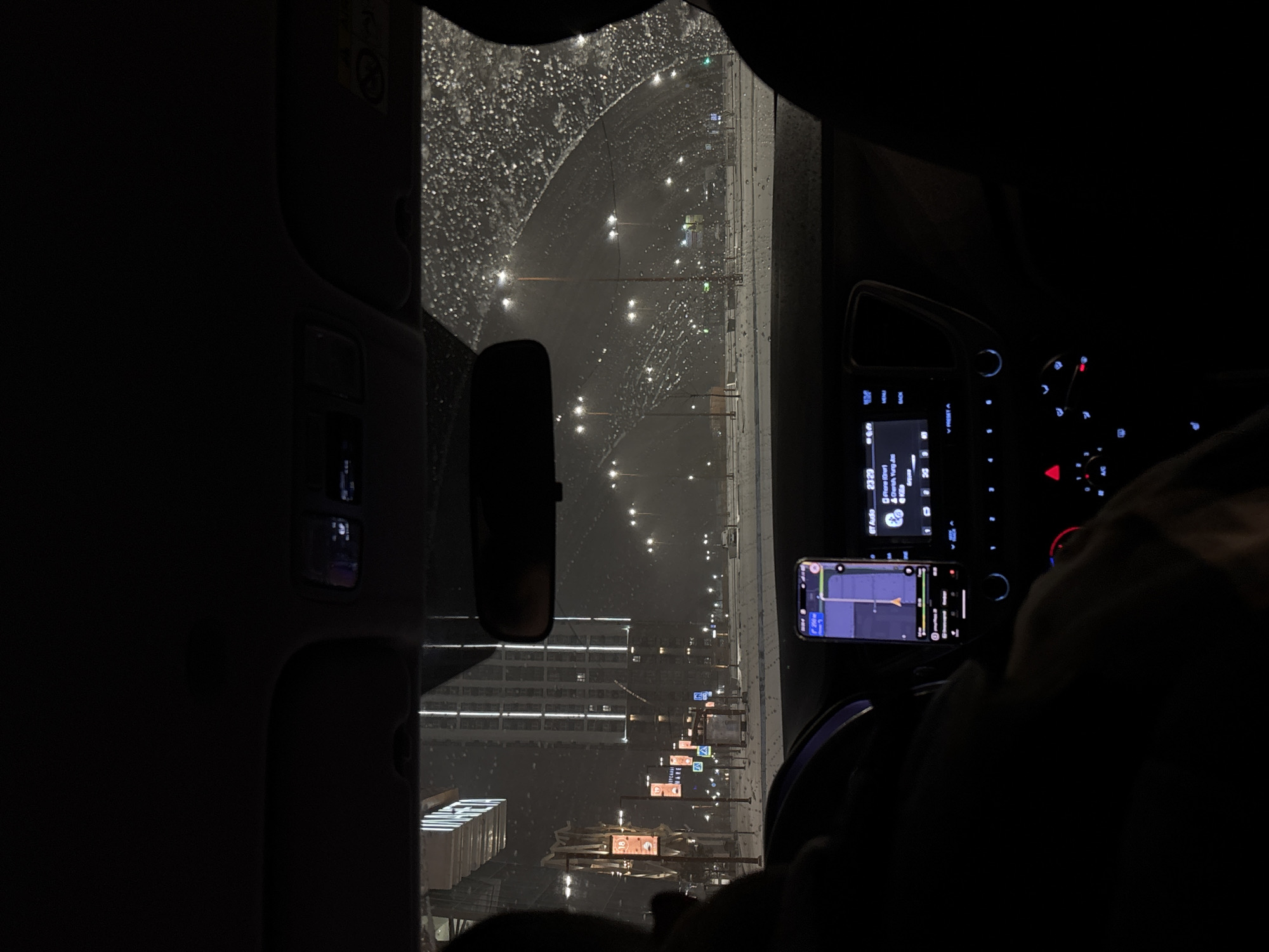 The image depicts a view from inside a car at night in Perm, Russia, during a rainstorm. The windshield is covered in raindrops. The street is visible through the windshield, showing city lights and building silhouettes. The dashboard of the car is visible, including a phone mounted with a navigation app running. The car's radio is illuminated. A rearview mirror is visible. A person's arm and shoulder are visible in the passenger seat. The city street is lined with street lights and building lights. Traffic signs are visible.