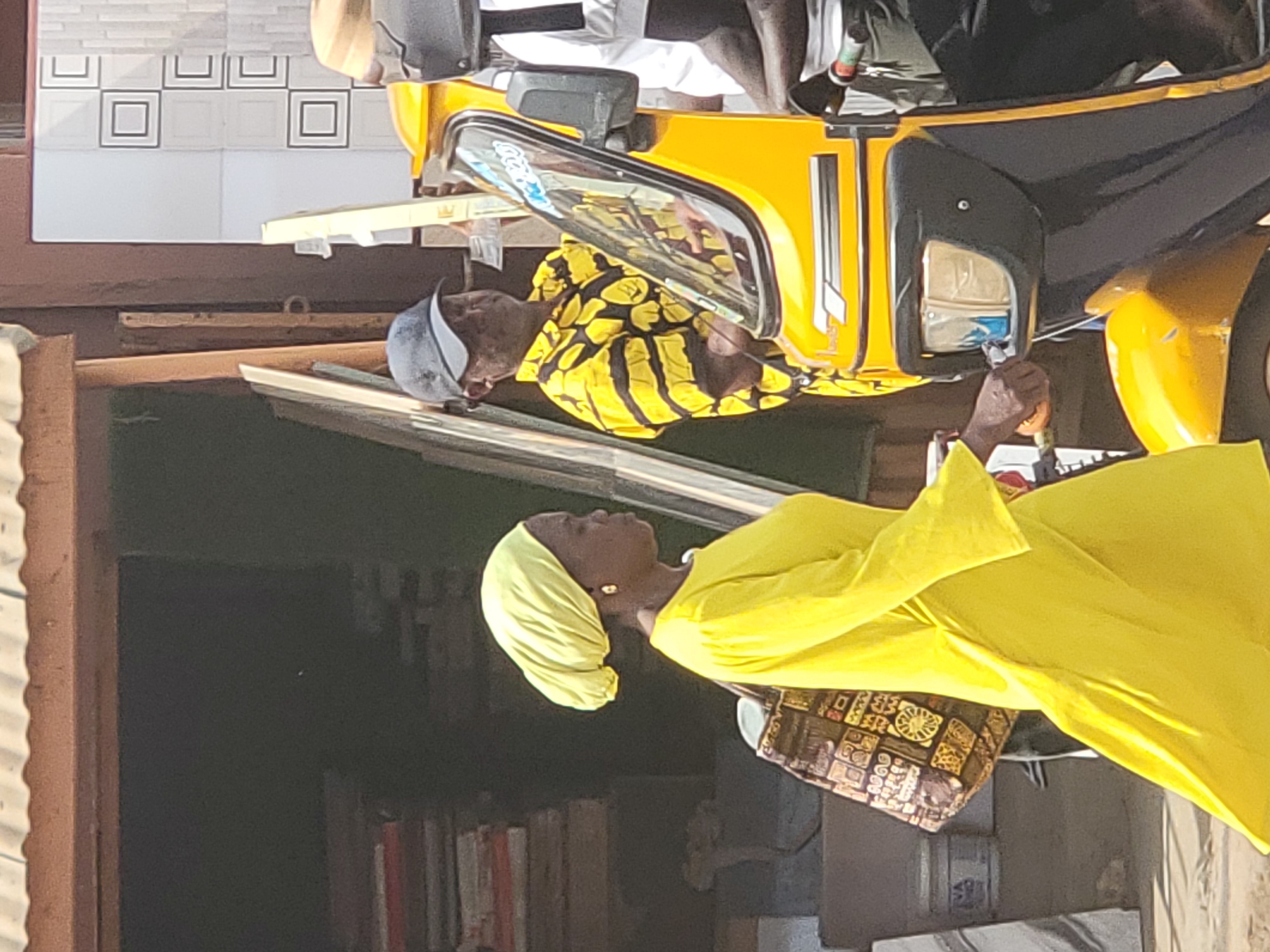 In the city of Maiduguri, Nigeria, a woman in a yellow dress and headscarf stands near a yellow and black auto rickshaw. An older man wearing a hat and yellow patterned shirt looks inside the vehicle. Another person, visible only from the torso up, stands in the auto rickshaw.