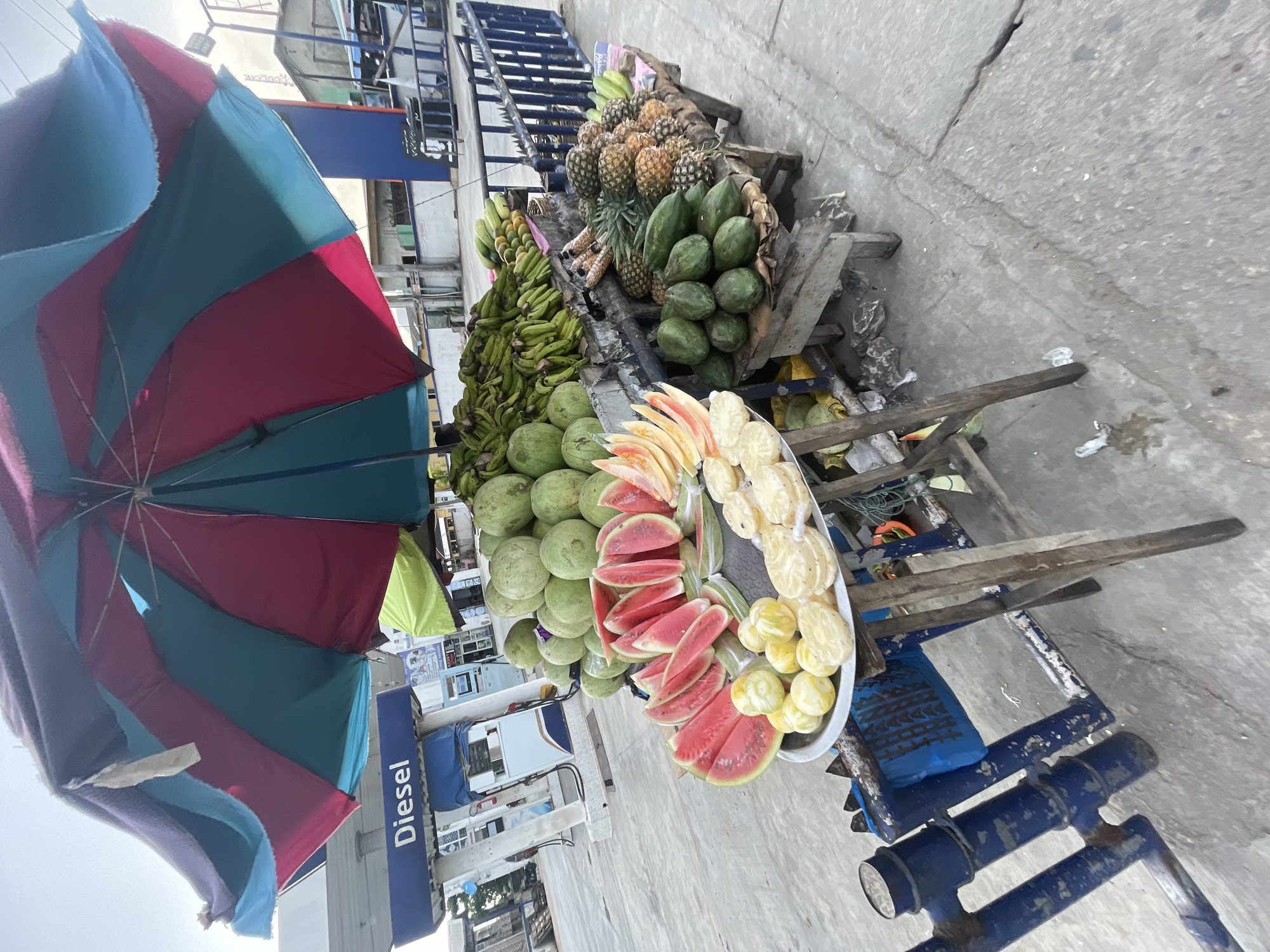 The image displays a vibrant outdoor fruit stall, likely situated on a paved roadside or market area, operating under the partial shade of a large, colorful umbrella. The umbrella features segments of red, blue/teal, purple, and light green fabric.
The fruit display is arranged on a rustic wooden cart with blue painted metal supports. A wide variety of fresh fruits are prominently featured. To the left, numerous bunches of green, unripe bananas are piled high. Adjacent to them are stacks of large, green, spherical fruits, which could be various types of melons or gourds. Further along the cart, a substantial quantity of pineapples are stacked, accompanied by several green, unripe papayas. The foreground of the stall showcases a white tray holding an assortment of neatly sliced fruits, including bright red watermelon slices and pale yellow or light green slices, possibly of another melon or citrus fruit, prepared for immediate consumption.
The ground beneath and around the cart is made of concrete or asphalt, showing some cracks and minor debris. In the background, various buildings are visible, and a notable blue sign with the word "Diesel" written in white text can be clearly seen, indicating the proximity of a gas station or fuel depot. The lighting suggests it is daytime, with a bright but possibly slightly overcast sky. No people are visible within the frame, but the organized display of goods implies an active vendor. The scene depicts a typical informal market setup, consistent with the provided location context of Ado, Nigeria.