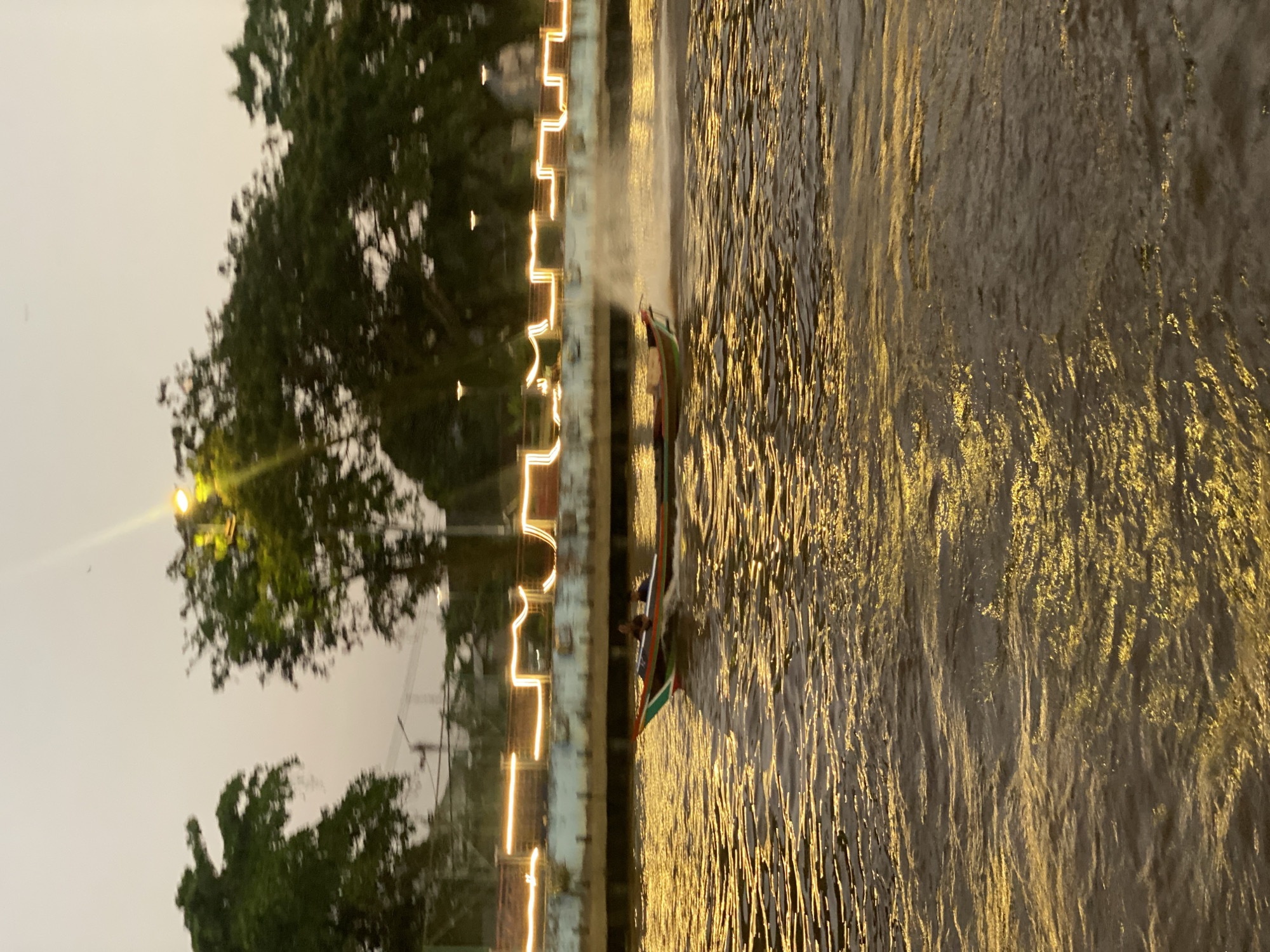 A long, narrow boat with two people onboard travels at speed down a murky river, leaving a spray of water in its wake. The boat has a colorful trim along its sides, with red, green, and white details. The river's surface is illuminated by a warm, golden light, reflecting the setting sun or artificial lighting from the embankment. Along the riverbank, a concrete structure is visible, adorned with a string of yellow lights that create a festive atmosphere. Trees with dense foliage line the top of the embankment, and in the distance, more lights and structures can be seen, suggesting an urban setting. This scene captures the essence of Banjarmasin, Indonesia, a city renowned for its riverine landscape and its vibrant floating markets, often referred to as the "City of a Thousand Rivers." The overall impression is one of evening activity on the water, with a blend of natural beauty and urban illumination.