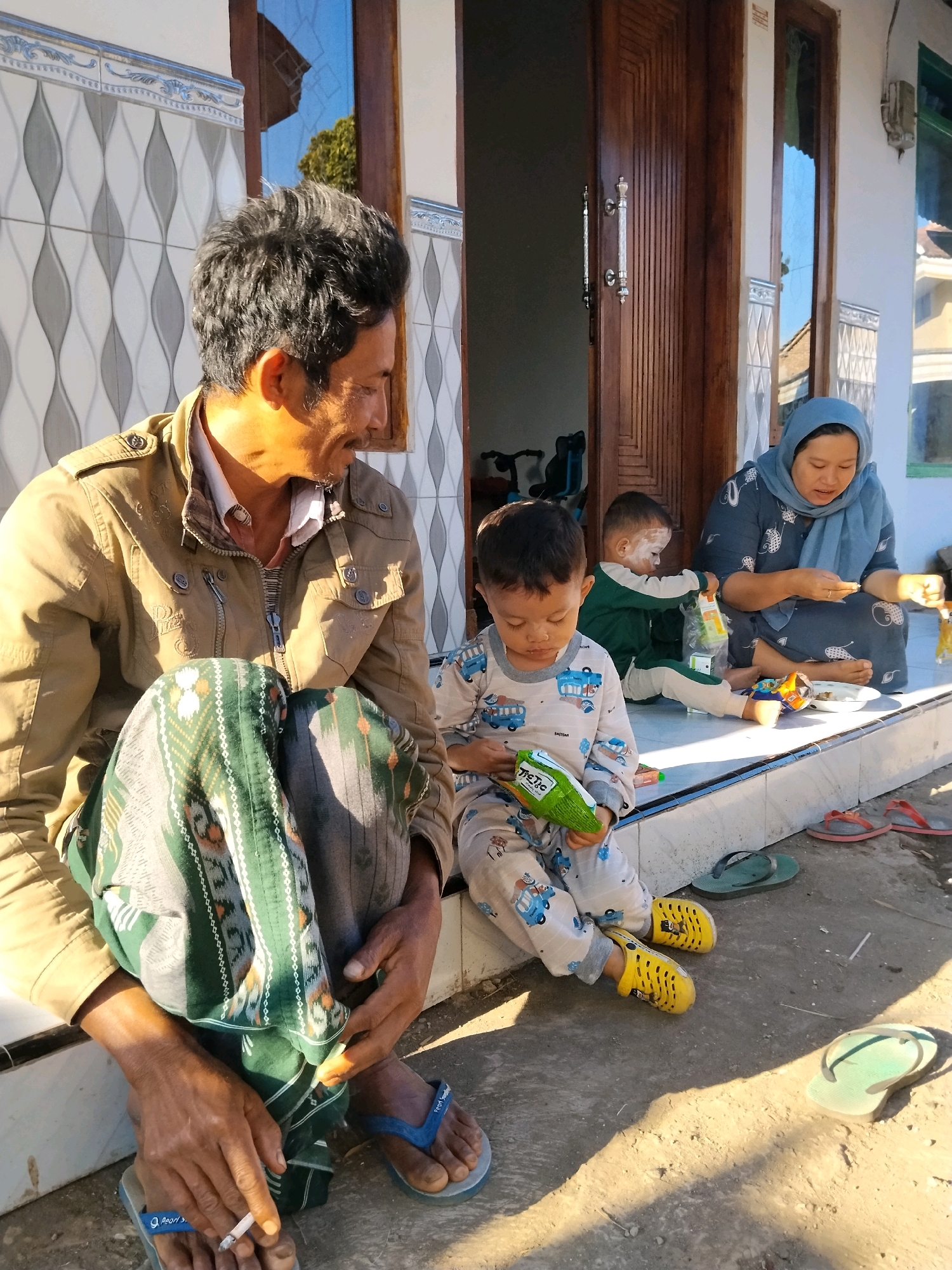 In front of a house in Panggungan, Indonesia, an adult male sits on a porch with his legs crossed, holding a lit cigarette between his fingers. He wears a tan jacket over a collared shirt and a green and white sarong. To his right, a young boy in light-colored pajamas patterned with cartoon vehicles sits on the porch and holds a green snack bag. Further to the right, a toddler wearing a green and white outfit sits and appears to be reaching for a snack bag held by a woman. The woman, wearing a blue hijab and a blue patterned dress, sits on the porch and holds a small object in her hands. The porch has a tiled border and the ground in front is dirt. Several pairs of flip-flops are scattered on the ground. The background shows a wooden door and white walls of the house.