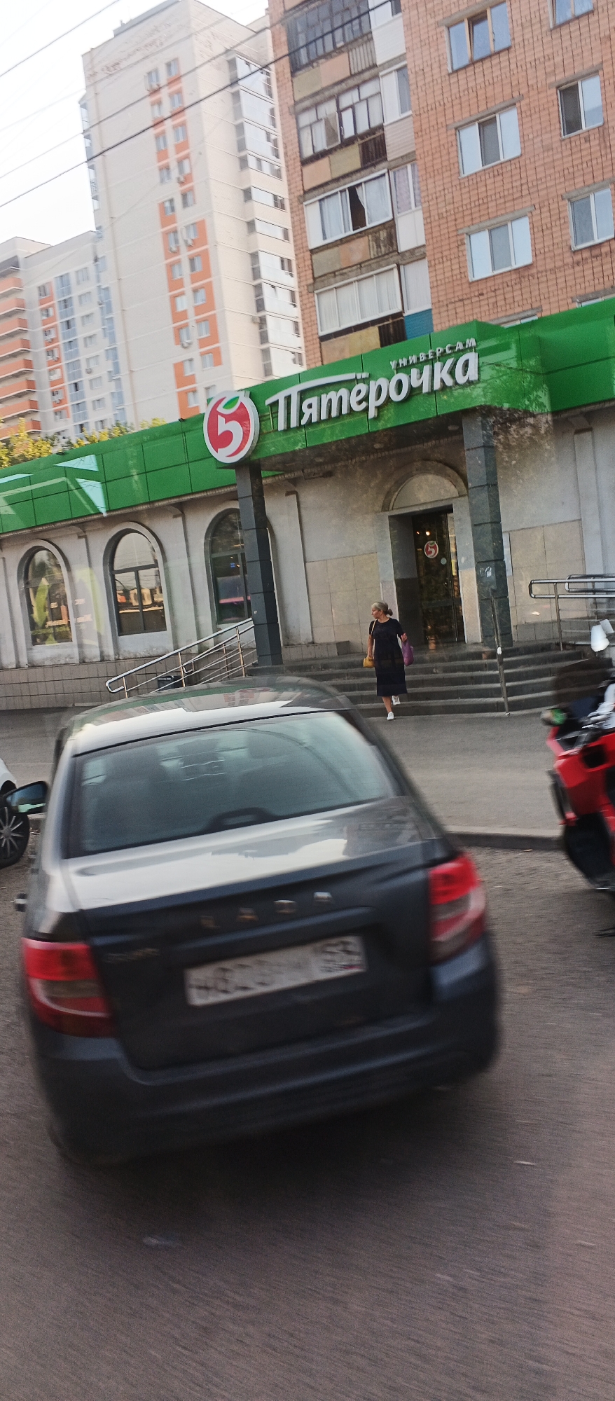 The image captures a street-level view of a Pyaterochka supermarket in Izhevsk, Russia. The facade of the supermarket is marked by its prominent green awning with the Pyaterochka logo, a red circle with a white number 5. Above the awning, a multi-story building with a brick facade and numerous windows is visible. To the left, another tall residential building with a lighter facade and orange accents is partially visible.
In the foreground, the rear of a dark-colored sedan, identified by the LADA emblem and a Russian license plate, is prominently featured. The car appears to be moving or has recently stopped, indicated by motion blur. A red motorcycle, also blurred due to motion, is parked to the right of the car.
On the steps leading into the supermarket, a woman in a dark dress and white shoes is walking. She carries a yellow purse and a shopping bag. The environment suggests an urban setting with a paved road and sidewalk area in front of the commercial establishment. The overall lighting indicates it is daytime, possibly late afternoon or early evening, given the shadows and the quality of light.