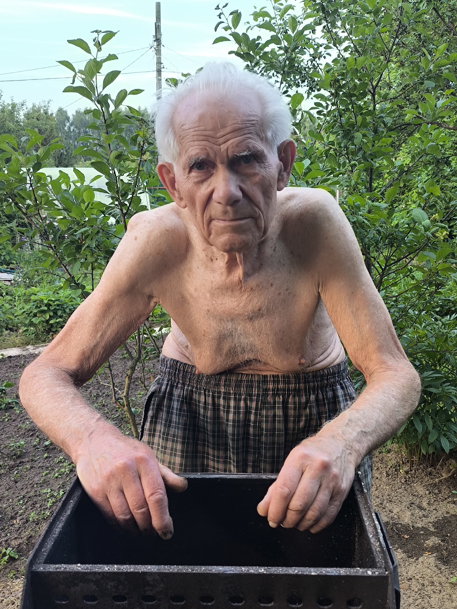 A shirtless elderly man with white hair, wearing plaid shorts, stands outdoors in front of a metal grill. He is leaning forward with his hands on the edge of the grill. The background consists of green foliage and trees, with a utility pole visible in the distance. The ground appears to be dirt.