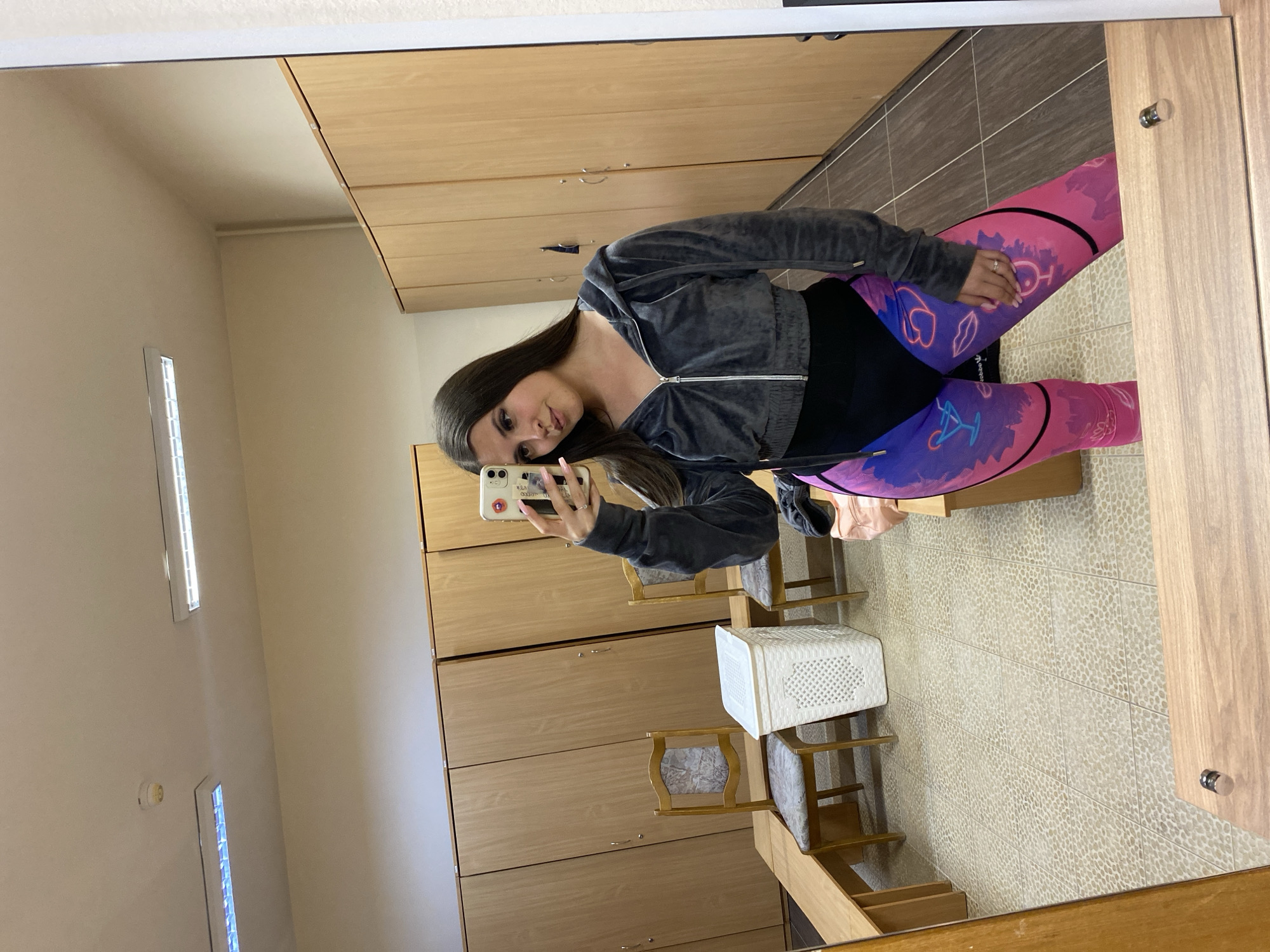 A young woman is captured in a mirror selfie, posing in what appears to be a changing room or locker room. She has long, dark brown hair and is wearing a dark gray, cropped zip-up hoodie over a black top. Her leggings are a vibrant mix of pink and purple, adorned with abstract, neon-style graphics including lips, martini glasses, and other stylized shapes. She wears a ring on her left ring finger and has long, light-colored nails. She holds a white-cased smartphone in her left hand to take the picture, displaying a neutral expression with slightly pursed lips.
The setting features rows of light-toned wooden lockers with metal handles, extending across the background. The floor is covered with light-colored, textured tiles. In the foreground, near the bottom of the mirror's reflection, are two wooden chairs with upholstered seats and a white, rectangular plastic woven basket. The room is brightly illuminated by long, rectangular ceiling lights, one of which is visible in the upper part of the reflection. The image itself is rotated 90 degrees clockwise. There is no clearly legible text visible.
