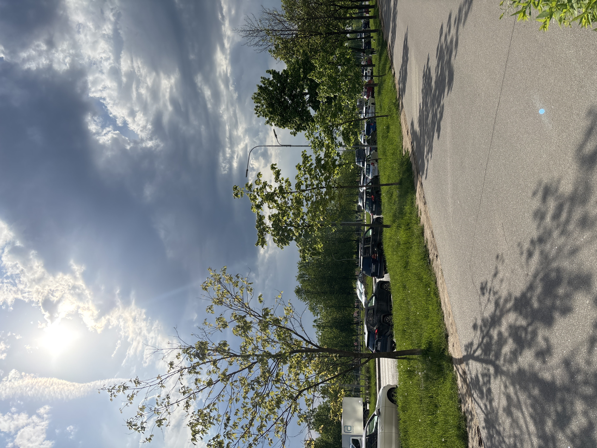 The image shows a street in an urban area, possibly a parking lot or roadside area, in Unknown, Russia. A row of parked cars lines the right side of the image. A paved road occupies the right side, with shadows cast by trees. A grassy area separates the road from the parked cars. The sky is visible in the background, with a mix of clouds and sun. The general environment suggests an outdoor scene during daylight. A light pole is visible among the trees. The image is rotated 90 degrees counterclockwise.