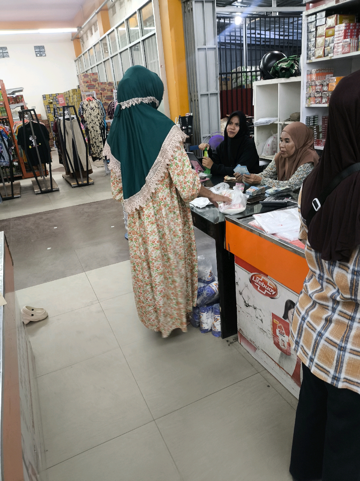 The image shows a retail environment, likely a convenience store or small supermarket, in Posso, Indonesia. Three women are visible, two working behind a counter and one in front, seemingly making a purchase. The woman in the foreground is wearing a floral print dress and a green hijab with lace trim. She is reaching towards a bag of items on the counter. Behind the counter, one woman in a black hijab is holding a bottle and looking towards the customer, while another woman in a brown hijab is counting money. Shelves stocked with various goods, including what appear to be packaged food items and personal care products like Lifebuoy, are visible in the background. A calculator is also present on the counter. The floor is tiled, and clothing racks with apparel are visible to the left of the scene.