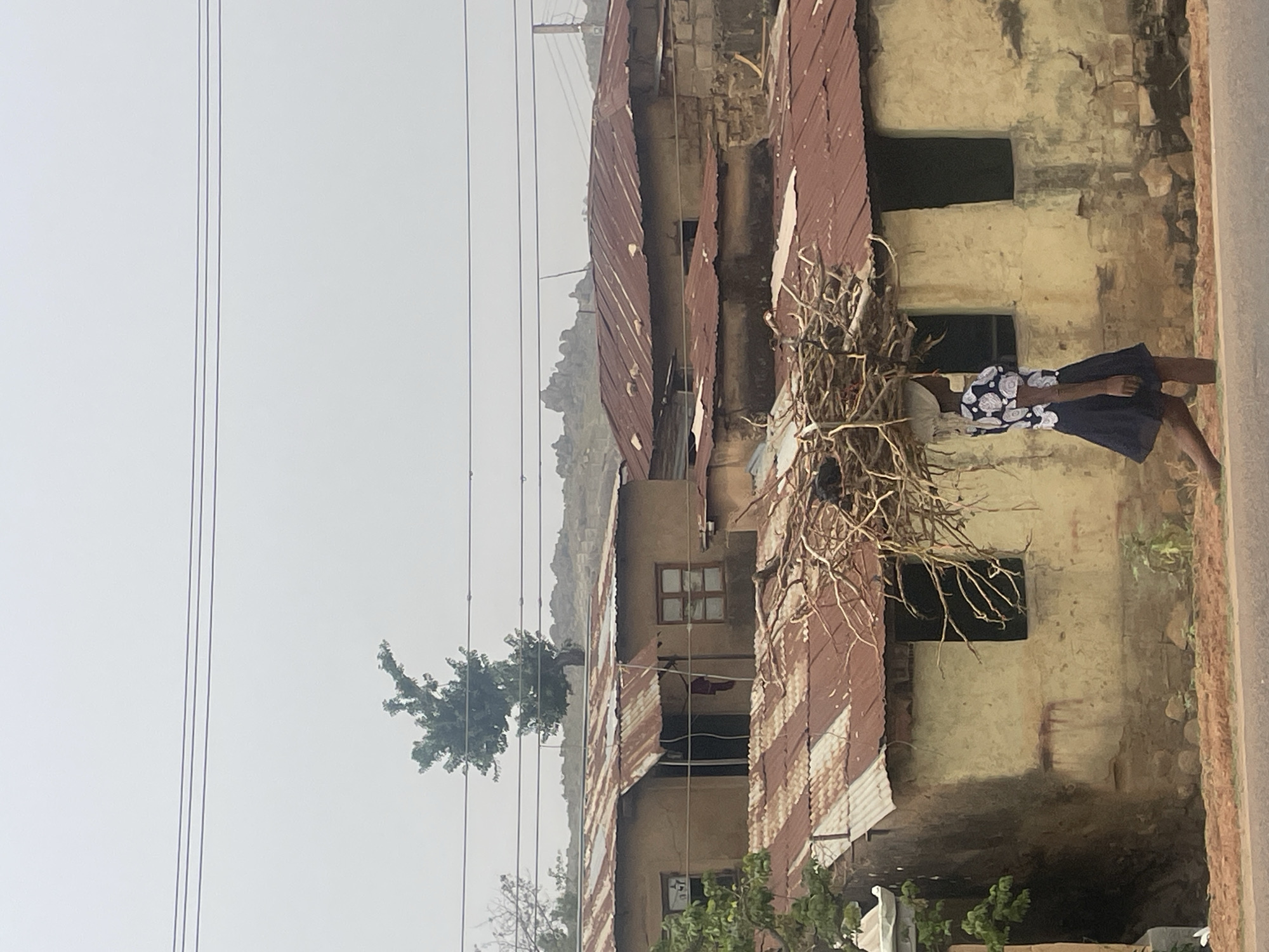 A young woman carries a bundle of branches near a dilapidated building in a residential area of Jos, Nigeria. The structure has a rusty corrugated metal roof and weathered, yellow-beige walls with dark, rectangular openings. Overhead power lines stretch across a hazy sky. Hills are visible in the background.