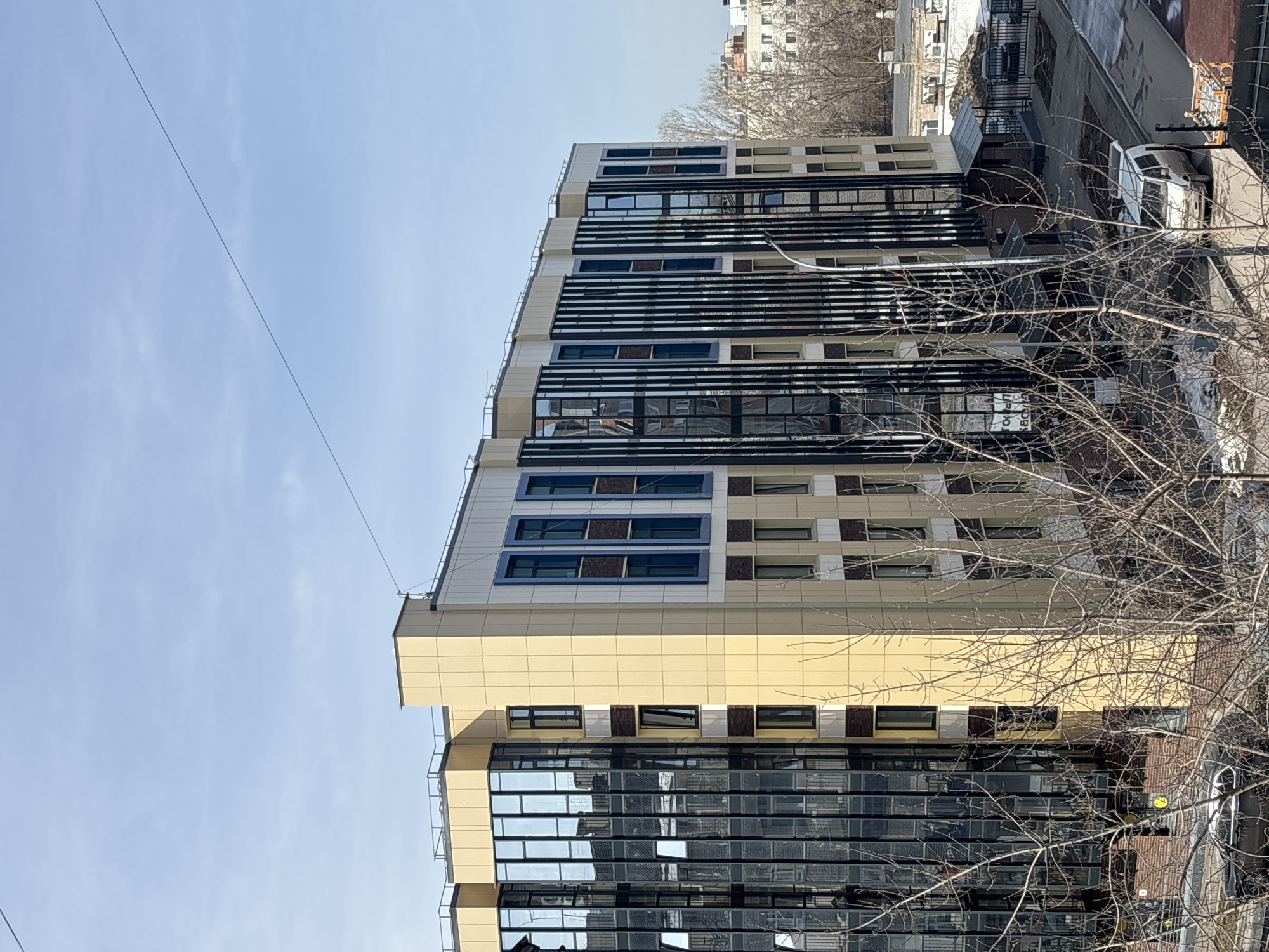 The image shows the facade of a modern building in Perm, Russia. The building has a multi-tiered design with sections featuring large glass windows, beige paneling, and sections with a pattern of smaller windows. A set of black, horizontal slats partially covers a section of the building. A few bare trees are in the foreground, partially obscuring the lower right side of the building. A utility line runs across the upper left portion of the image. In the background, parts of other buildings and a white van are visible. The sky is pale blue.