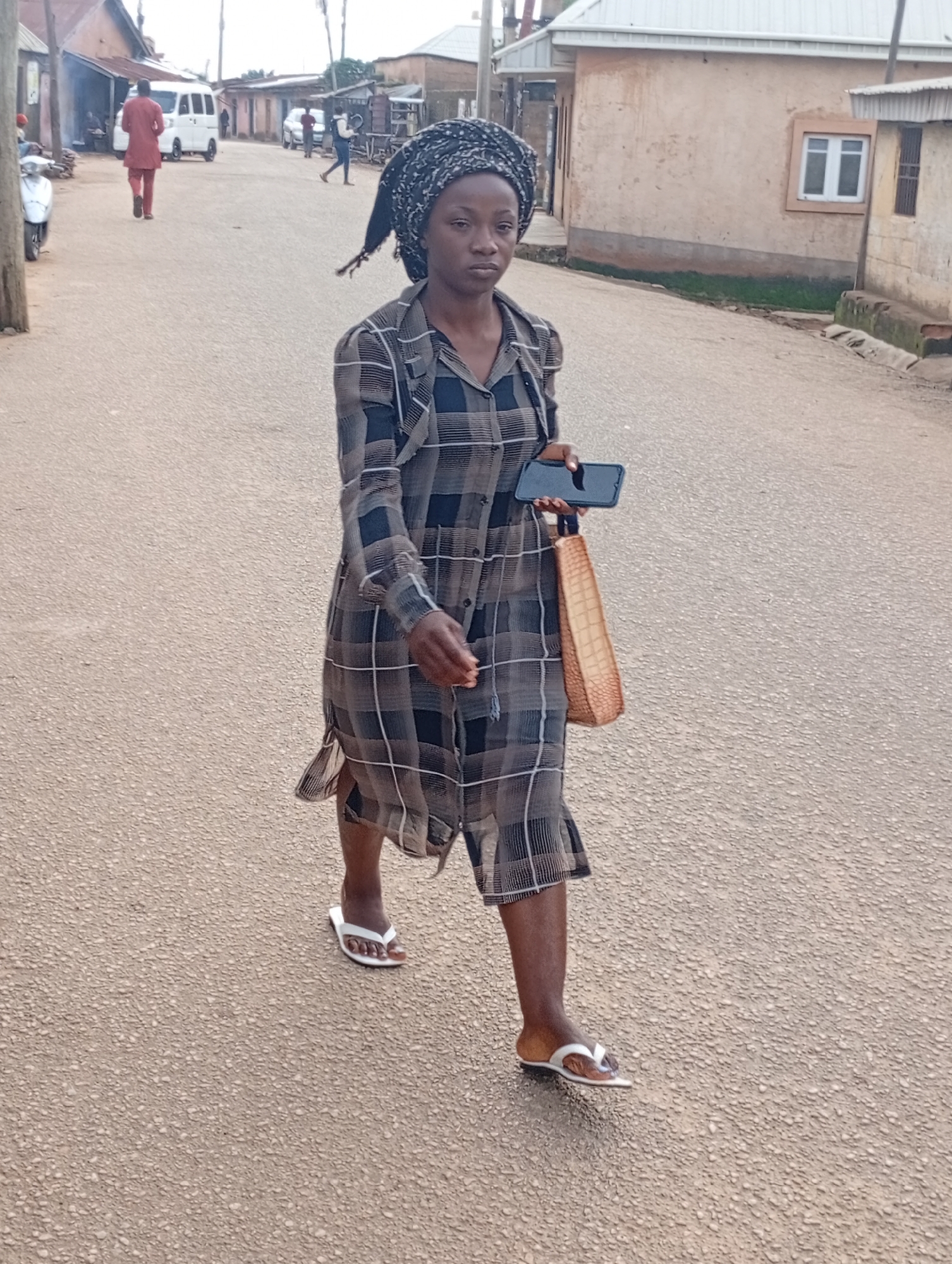 A woman wearing a headscarf and a checkered dress walks on a paved street in a village. She carries a smartphone and a woven bag. Other people and vehicles are visible in the background, including a white van and a motorcycle. Buildings with corrugated iron roofs line the street.