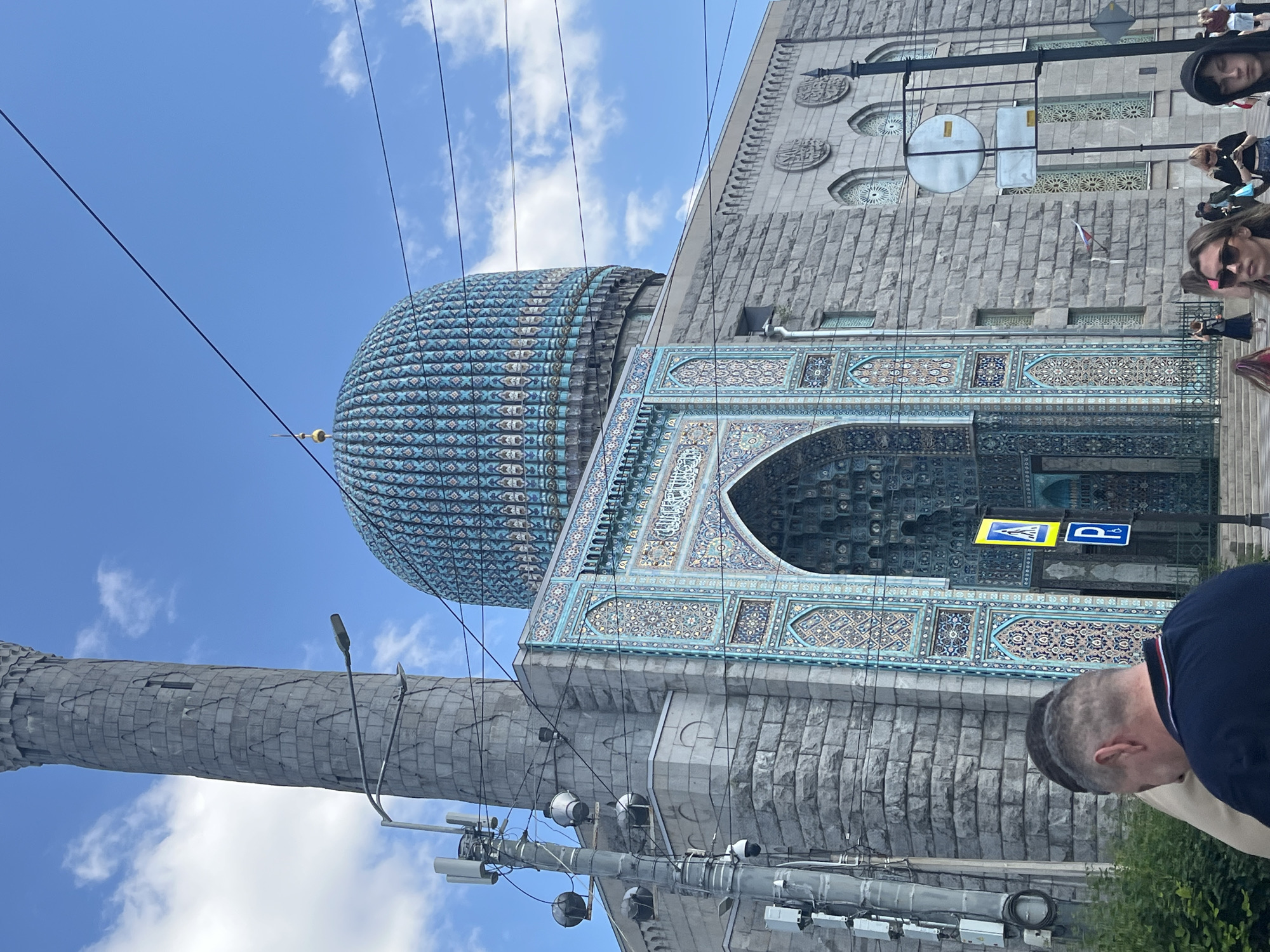 The image shows the Saint Petersburg Mosque, a large mosque located in Saint Petersburg, Russia. The mosque features a prominent blue dome and a tall minaret. The building is constructed of grey stone and is elaborately decorated with intricate blue and white tilework, particularly around the entrance. A busy street scene is visible in the foreground, with several people walking and standing. Traffic lights and street signs are also present. The sky is blue with scattered clouds, indicating daytime. The camera angle is from street level, looking up at the mosque.