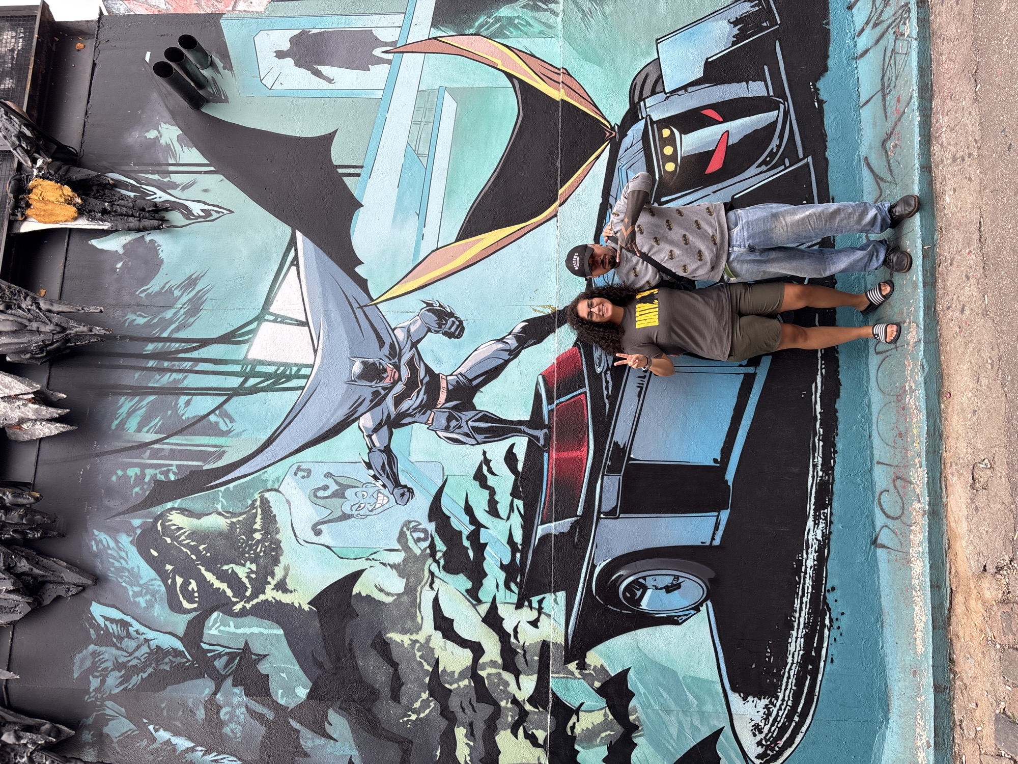A color photograph displays a vibrant mural featuring the character Batman in action, taken at an outdoor location in São Paulo, Brazil. The mural depicts Batman, rendered in a comic book style, leaping in front of a large vehicle with a bat-like design. The background includes elements of Gotham City, with stylized buildings, bats, and smoke. To the right of the mural, two individuals are posing. The person closer to the mural wears a gray T-shirt with a Batman logo, green shorts, and black and white striped sandals. They have dark curly hair and are holding up two fingers in a peace sign. The other person is wearing a grey sweatshirt with a Batman print, blue jeans, and black shoes, along with a dark baseball cap, also flashing a peace sign. The mural itself is textured and slightly damaged. The location appears to be an alleyway or a similar urban setting with exposed brick and graffiti-like markings on the wall.