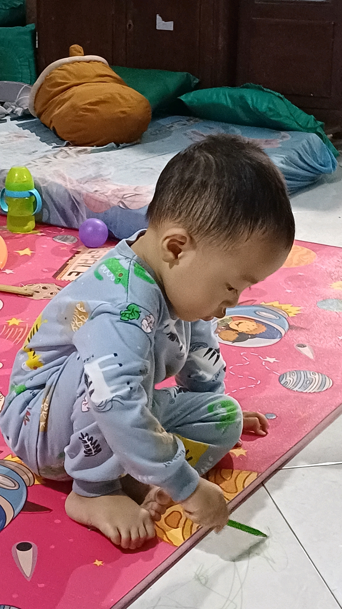 In Wonokerso, Indonesia, a young child is sitting on a pink playmat in a room. The child is wearing blue pajamas with animal prints and is focused on something on the mat. Nearby, there are several green pillows and a large, orange, acorn-shaped pillow. A blue and green sippy cup and a purple ball are also visible on the mat. The playmat has colorful graphics, including planets and cartoon characters. The flooring in the room appears to be white tiles.