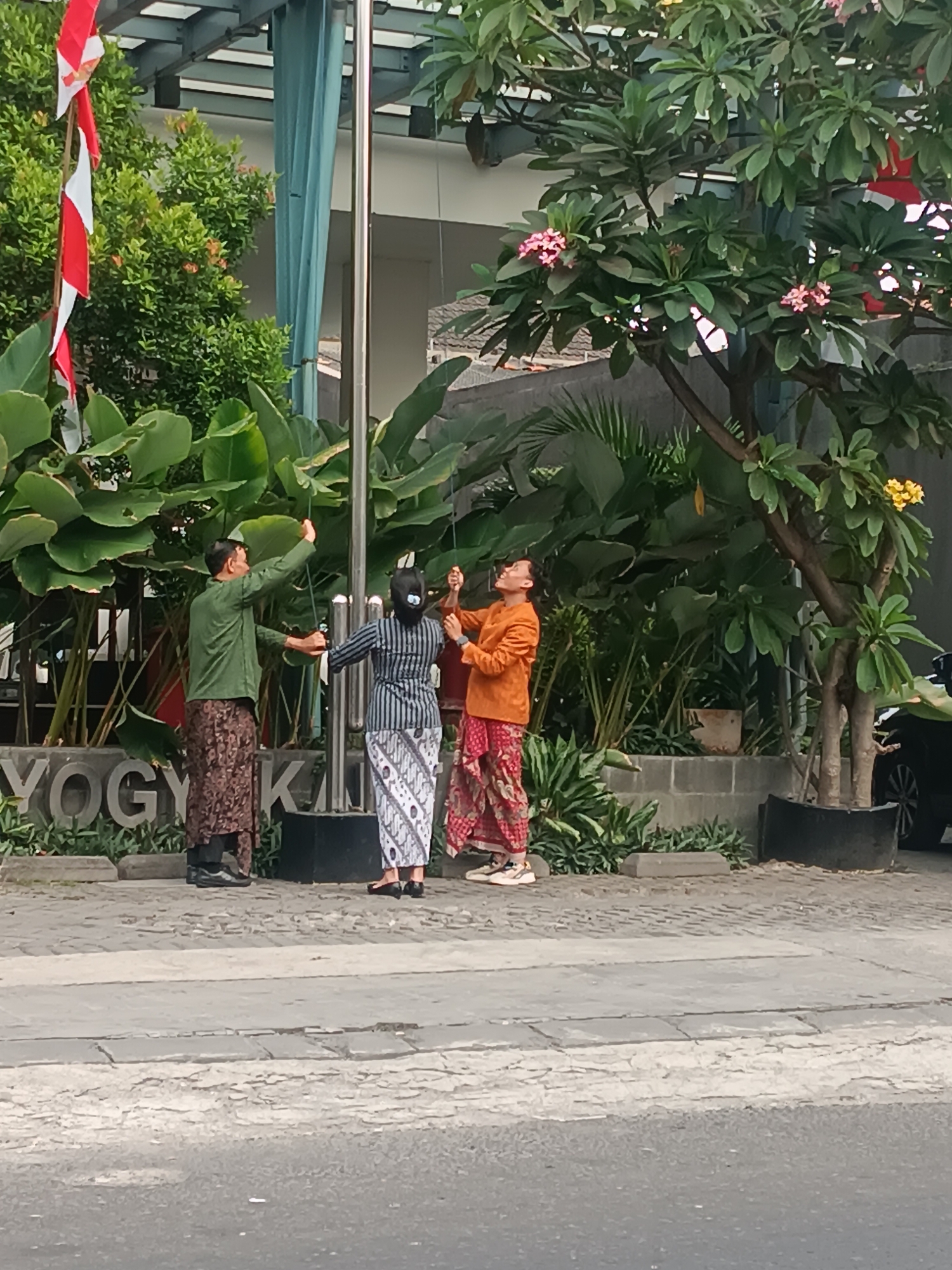 In Yogyakarta, Indonesia, three individuals are captured in a moment of flag-raising. The scene unfolds on a paved outdoor area, likely a public space given the prominent "YOGYAKARTA" lettering in large, three-dimensional gray characters behind the individuals. To the left, a man in a green long-sleeved shirt and a batik sarong is seen reaching upwards, holding a rope. Beside him, a person in a striped black and white outfit, also wearing a batik sarong, mirrors his action, grasping another rope. To the right, a third individual, dressed in an orange long-sleeved shirt and a red and white patterned batik sarong, looks up and holds a rope. They appear to be positioned at the base of a flagpole, suggested by the ropes and their upward gazes. The background features lush green foliage, including large-leafed plants and a tree with pink flowers, hinting at a tropical climate. A portion of a building with a light blue curtain is visible, along with a black car parked on the right side of the frame. The overall atmosphere suggests a formal ceremony or event.