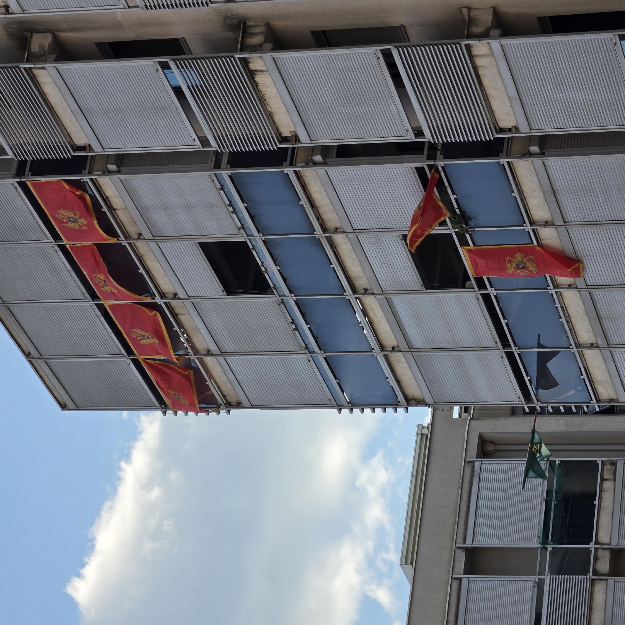 The image is an upward-facing shot of two buildings in Podgorica, Montenegro, under a blue sky with some clouds. The first building has a series of metal sunshades or louvers and glass panels, with red Montenegrin flags hanging from several windows. The second building is visible on the right side of the frame, featuring a green flag hanging on a balcony. The architectural style is modern, with the sunshades casting shadows. The image has a slightly disorienting perspective, seemingly taken from directly below the buildings.