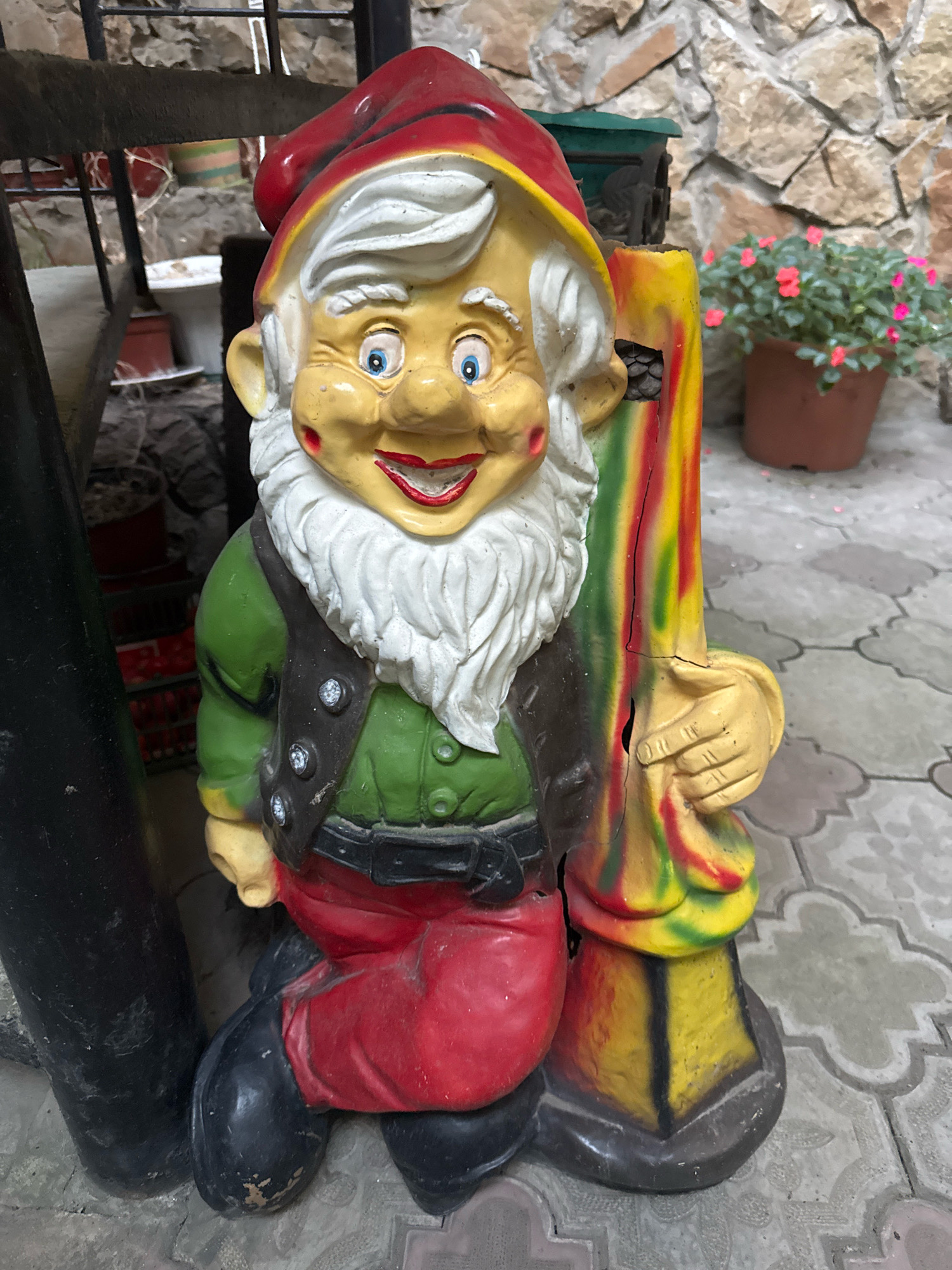 An old, weathered garden gnome statue stands on a patterned paved surface in Anapa, Russia. The gnome is made of painted material, likely concrete or plaster, and shows signs of wear and cracking. It is depicted with a red hat, a yellow face, white beard and eyebrows, blue eyes, and rosy cheeks. The gnome wears a green tunic over a red garment, a dark vest with decorative buttons, and black boots. It holds a colorful, cracked object in its right hand, painted with vertical stripes of red, yellow, and green. Behind the gnome, a stone wall and some potted plants are visible, including a terracotta pot with pink flowers. A metal railing and a dark, cylindrical object are also present in the background.