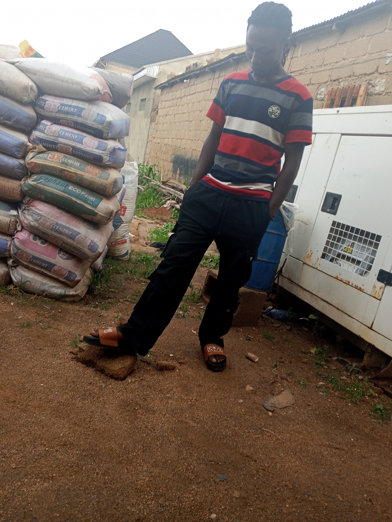 A young Black male with dark braided hair stands with his right foot on a small rock in what appears to be an outdoor construction or industrial area. He is wearing a horizontally striped t-shirt in blue, red, and grey, with a circular emblem on the left chest. He also wears dark, loose-fitting cargo pants and brown sandals with an orange strap. His right hand is in his pocket.
To his left, a large stack of bagged cement is visible, with various brands and colors, including "DANCOTE CEMENT" and "Elephant ECOPlanet." Behind him, a large white generator unit is situated, with a blue barrel next to it. The ground is covered in reddish-brown dirt and gravel. In the background, a brick building with a dark pitched roof is present, along with some overgrown vegetation. The sky is overcast. The overall scene suggests a work in progress or storage area, likely in an urban setting like Jos, Nigeria, characterized by construction materials and equipment.