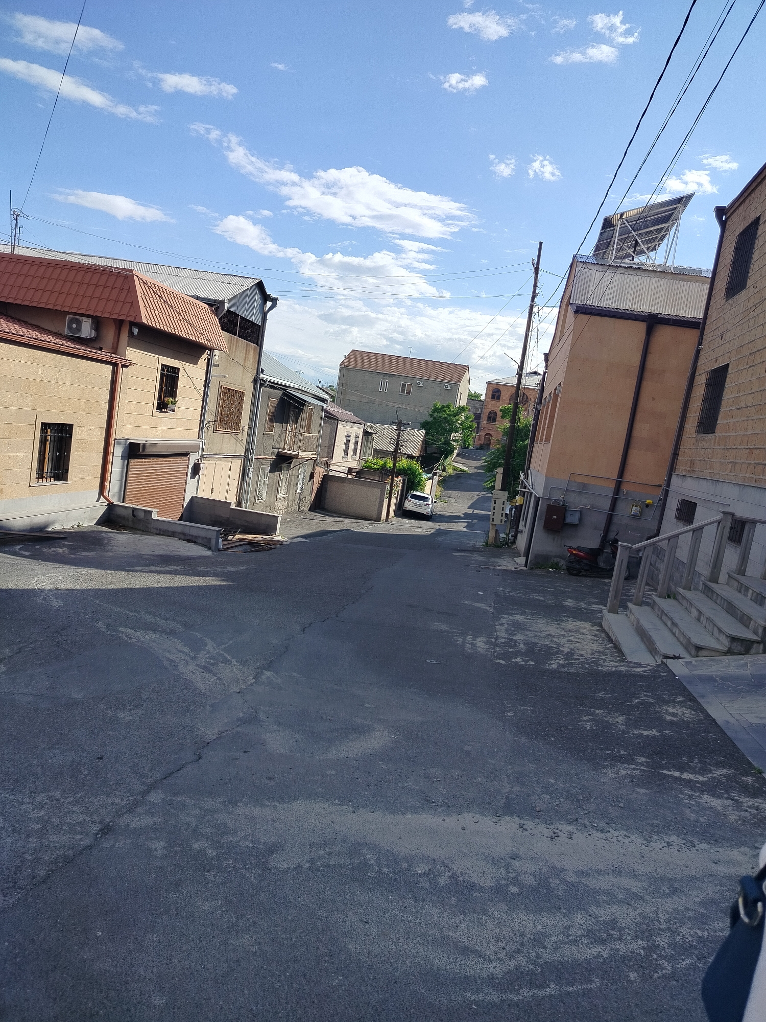 The image depicts a street scene in Yerevan, Armenia. The perspective is from street level, looking uphill along a paved road. The road surface is gray and shows signs of wear and repair. Buildings of varying heights and architectural styles line both sides of the street. The buildings are primarily constructed of stone or brick, with facades in shades of beige, brown, and gray. Some buildings have visible windows, some with grilles, and at least one has a visible balcony. Roofing materials vary, including red tile and corrugated metal. Utility lines are visible overhead, strung between buildings and poles. A few parked vehicles are seen further up the street. The sky is partly cloudy with a blue hue. Vegetation, including trees and shrubs, is sparsely present along the road. There are also some steps on the right.