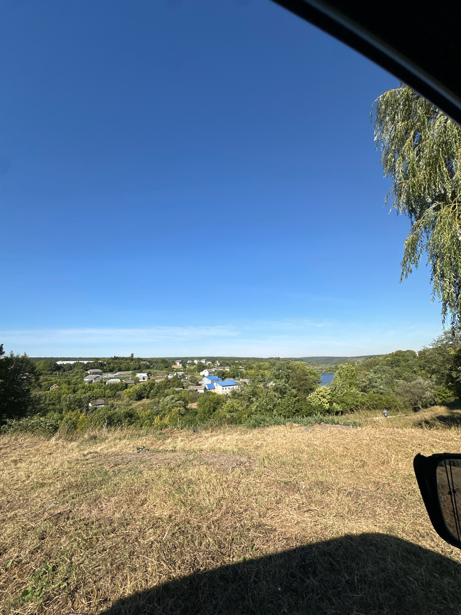 The image captures a wide-angle view of a rural landscape in Makiv, Ukraine, on a clear, sunny day. In the foreground, dry, golden-brown grass covers a hillside, casting a shadow from an unseen object on the right. A side mirror, likely from a vehicle, is visible on the lower right edge of the frame, reflecting a portion of the sky.
The middle ground reveals a cluster of houses with white and blue roofs, nestled amidst lush green trees and vegetation. A body of water, possibly a river or lake, is visible in the distance behind the houses, surrounded by rolling hills covered in dense foliage. A lone figure, appearing to be walking, is present on a path in the middle distance.
The upper portion of the image is dominated by a vast expanse of clear, bright blue sky. On the upper right, the drooping branches of a weeping willow tree are visible, partially framing the scene. The overall impression is one of a tranquil, sun-drenched rural setting.