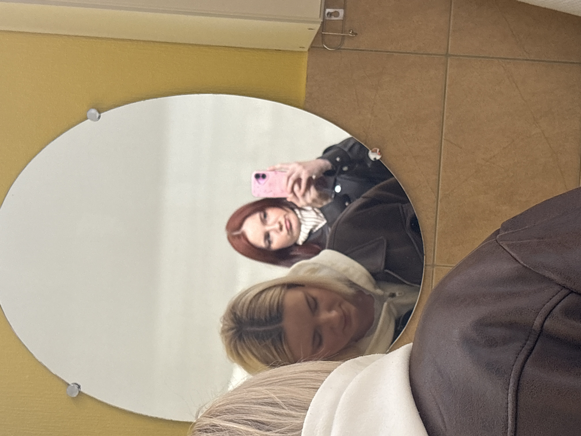 The image depicts a reflection in a circular mirror. The mirror is mounted on a yellow wall with visible hardware. The reflection captures two individuals: one with auburn hair holding a pink mobile phone, possibly taking a photo, and another with blonde hair resting their head on what appears to be a white fabric. A dark brown leather jacket is partially visible, belonging to an individual positioned outside the frame, closer to the viewer. The background suggests a bathroom setting with beige tiles. The location is Unknown, Unknown.