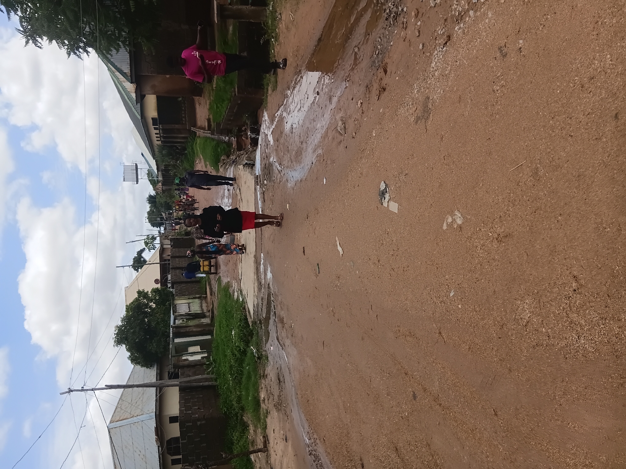 The image displays an unpaved, reddish-brown dirt road in a residential area of Jalingo, Nigeria, under a sky with scattered white clouds. The road surface is uneven, showing numerous tire tracks and evidence of recent water flow, including muddy streaks, shallow puddles, and damp patches. Small pieces of debris are scattered across the road.
On the left side of the road, a concrete block wall fronts a single-story building with a light-colored, corrugated metal roof. A wooden utility pole with overhead power lines extends vertically. In the mid-ground, a woman in a black top and red skirt, wearing sandals, walks towards the viewer, holding a mobile phone in both hands. Further behind her, a man in a dark shirt and trousers walks away from the viewer, and a small group of people are visible in the distant background.
On the right side of the road, a concrete drainage gutter, partially filled with muddy water, runs parallel to the road. Patches of green grass and sparse vegetation grow along the gutter. A man in a pink t-shirt and dark trousers stands facing partially left, holding a mobile phone. Behind him, a person in a patterned dress walks away from the viewer, pulling a two-wheeled handcart loaded with two yellow jerrycans. Further down this side, several buildings with corrugated metal roofs, some painted green, line the road. In the background, a white water tank on a raised metal frame is prominent.