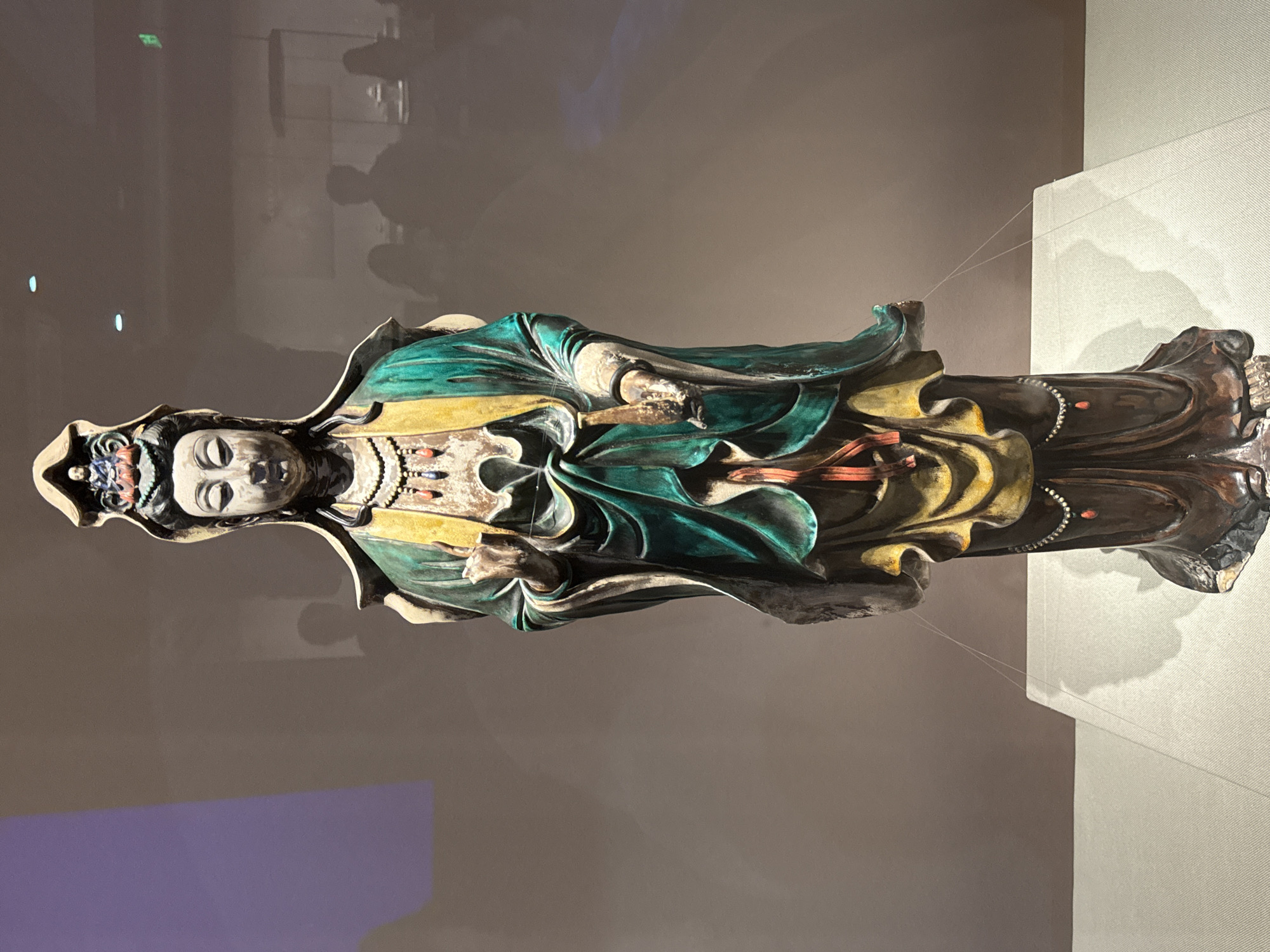 The image shows a colorful statue of a figure in a museum or exhibit. The statue is of a person wearing a flowing robe with intricate details and multiple colors, including green, yellow, red, and dark brown. The face of the statue is light-colored, with distinct facial features. The statue stands on what appears to be a base. Behind the statue, there's a wall with a light grey hue. Reflections of people can be seen on the wall, indicating the presence of visitors. The statue is indoors. Without specific information, it is impossible to confirm that the museum is located in Yuzhong District, China.