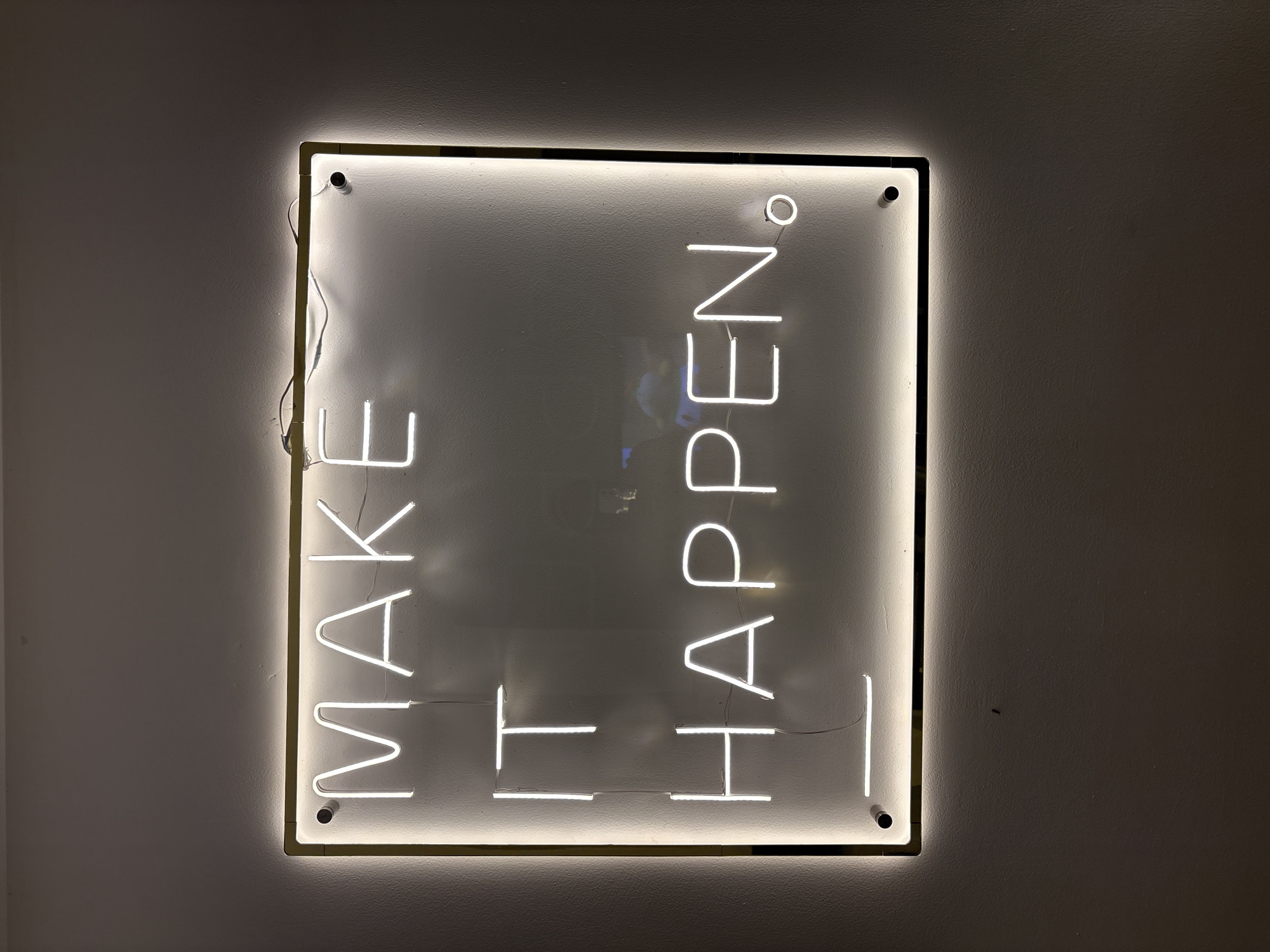 A square neon sign with the words "MAKE IT HAPPEN" vertically arranged hangs on a light-colored wall. The sign has a metallic frame with illuminated edges, and the lettering is in a bright white neon. The words are oriented in two columns, with "MAKE IT" on the left and "HAPPEN" on the right. The background of the sign is a slightly reflective white or light gray. The wall is plain with a subtle texture. The location is unknown.