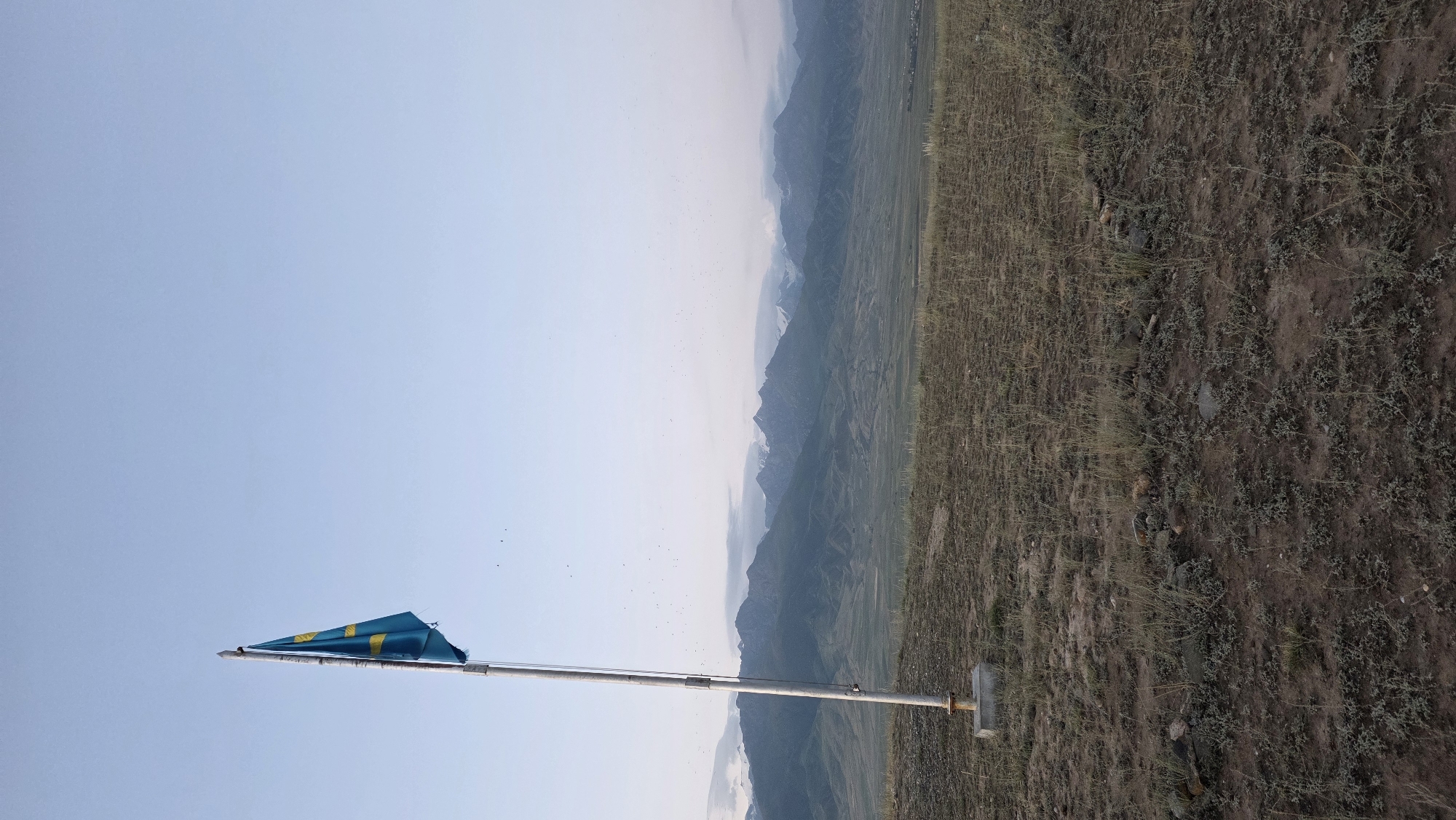 A Kazakhstan flag is seen on a flagpole to the left of the frame. The flag is blue with a gold sun and eagle. The flagpole is metal and stands on a concrete base. In the background, there are mountains under a clear sky with some clouds. The foreground is a grassy field with some rocks. The scene appears to be in a rural area.