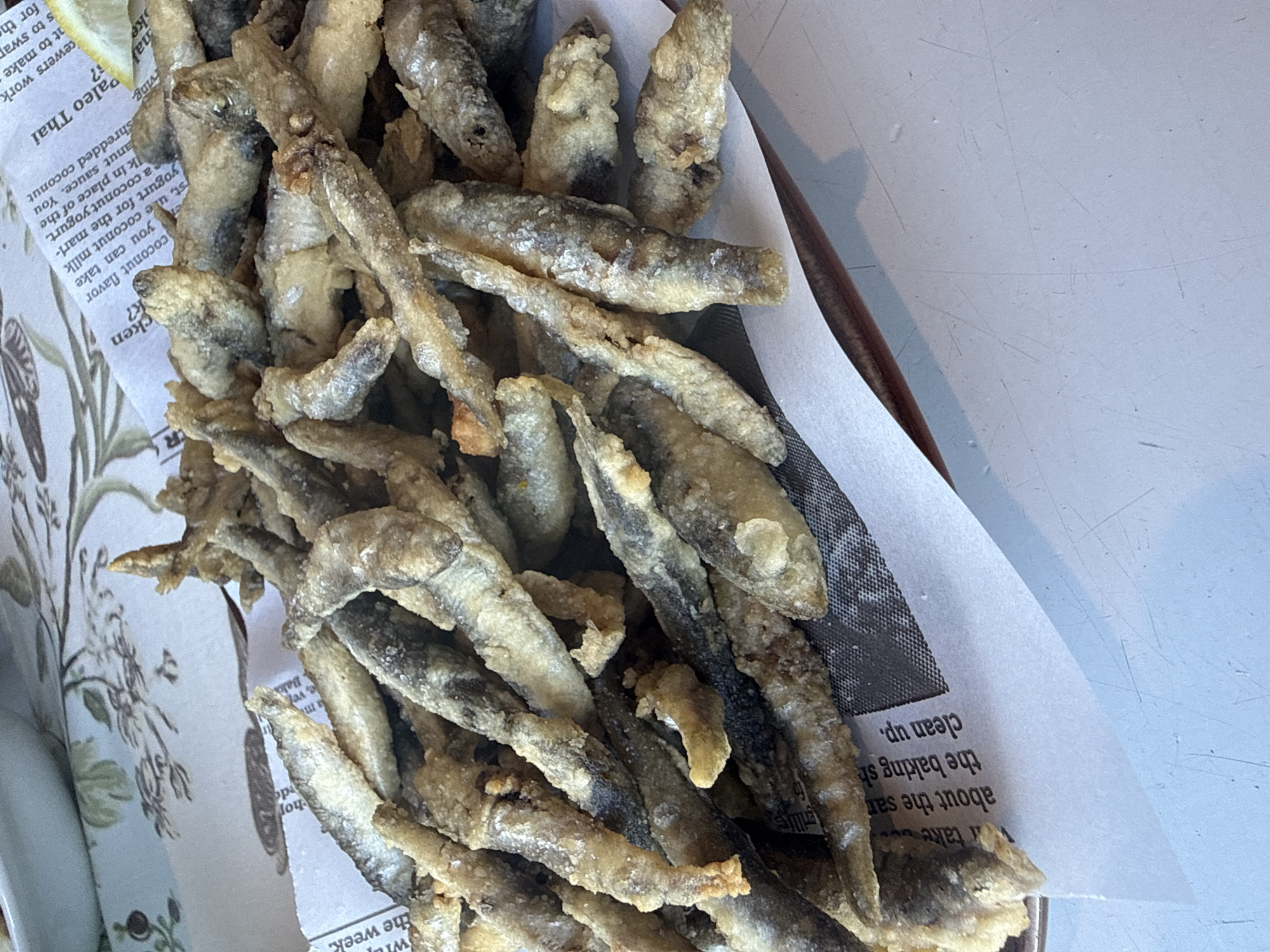 This is a top-down, close-up shot of a dish of small, fried fish served on top of newspaper. The fish are golden brown and appear crispy, with many of them whole and intact. They are piled generously in a basket lined with paper. The newspaper beneath the fish has printed text and images, with some of the text visible including "Paleo Thai", "coconut flavor", and "clean up.". The scene appears to be a casual meal setting. There are no people visible. The lighting is bright, suggesting it could be daytime, and the background is a light-colored, textured surface, possibly a table.