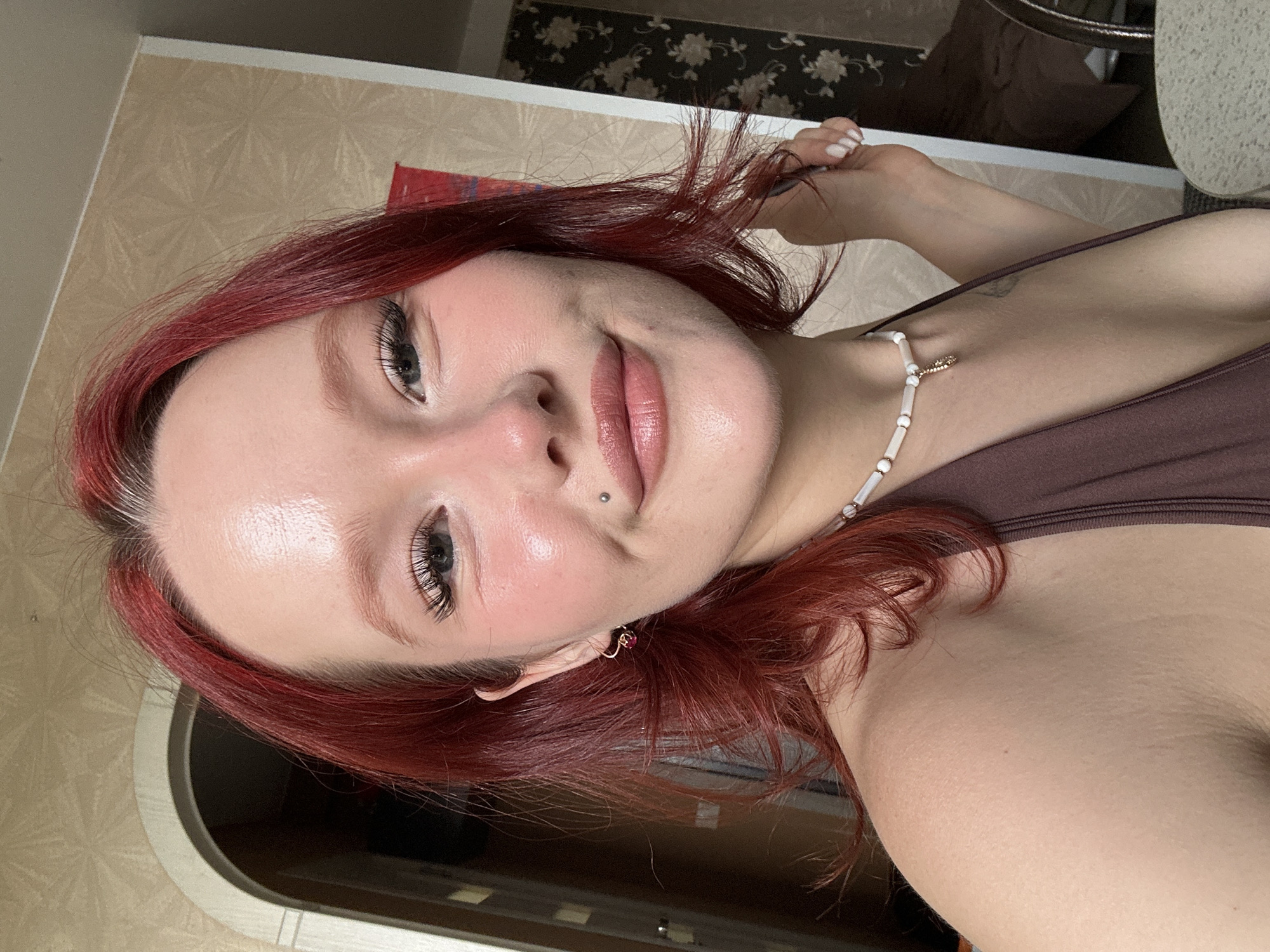 A young woman with vibrant red hair is taking a selfie in a room. She has fair skin with blush and highlighted cheekbones, light-colored eyebrows, and dramatic false eyelashes. A small silver stud is visible on her lower lip. She is wearing a brown, halter-neck top and a beaded necklace with a small pendant. Her left arm is slightly extended, with her hand partially visible, and her right hand is near her hair. The background appears to be an interior space with textured beige wallpaper and an arched doorway on the left.