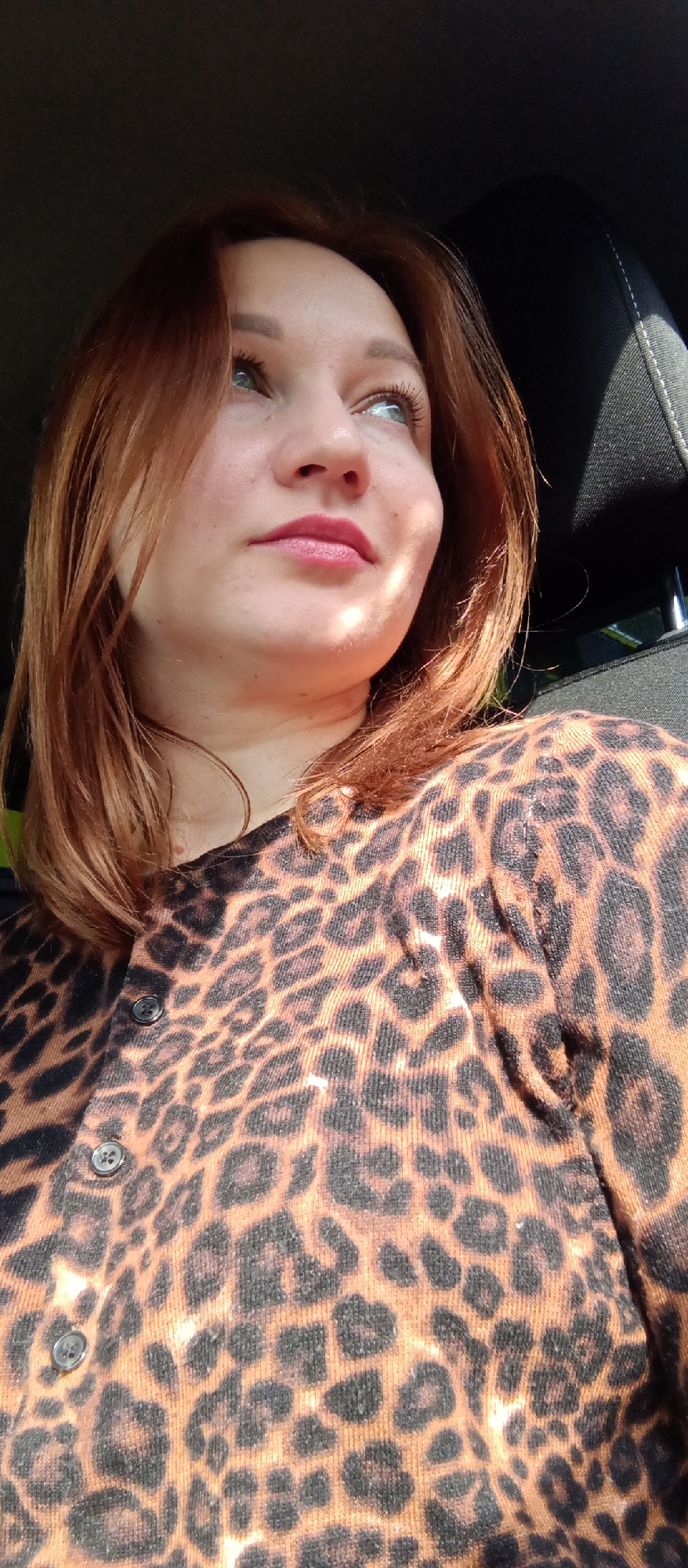 A woman with auburn hair is pictured from a low angle, looking upwards and slightly to the right. She is wearing a leopard print cardigan with buttons visible. The setting appears to be inside a car, evidenced by the black headrest visible in the background. The specific location within Unknown, Unknown is not discernible from the image.