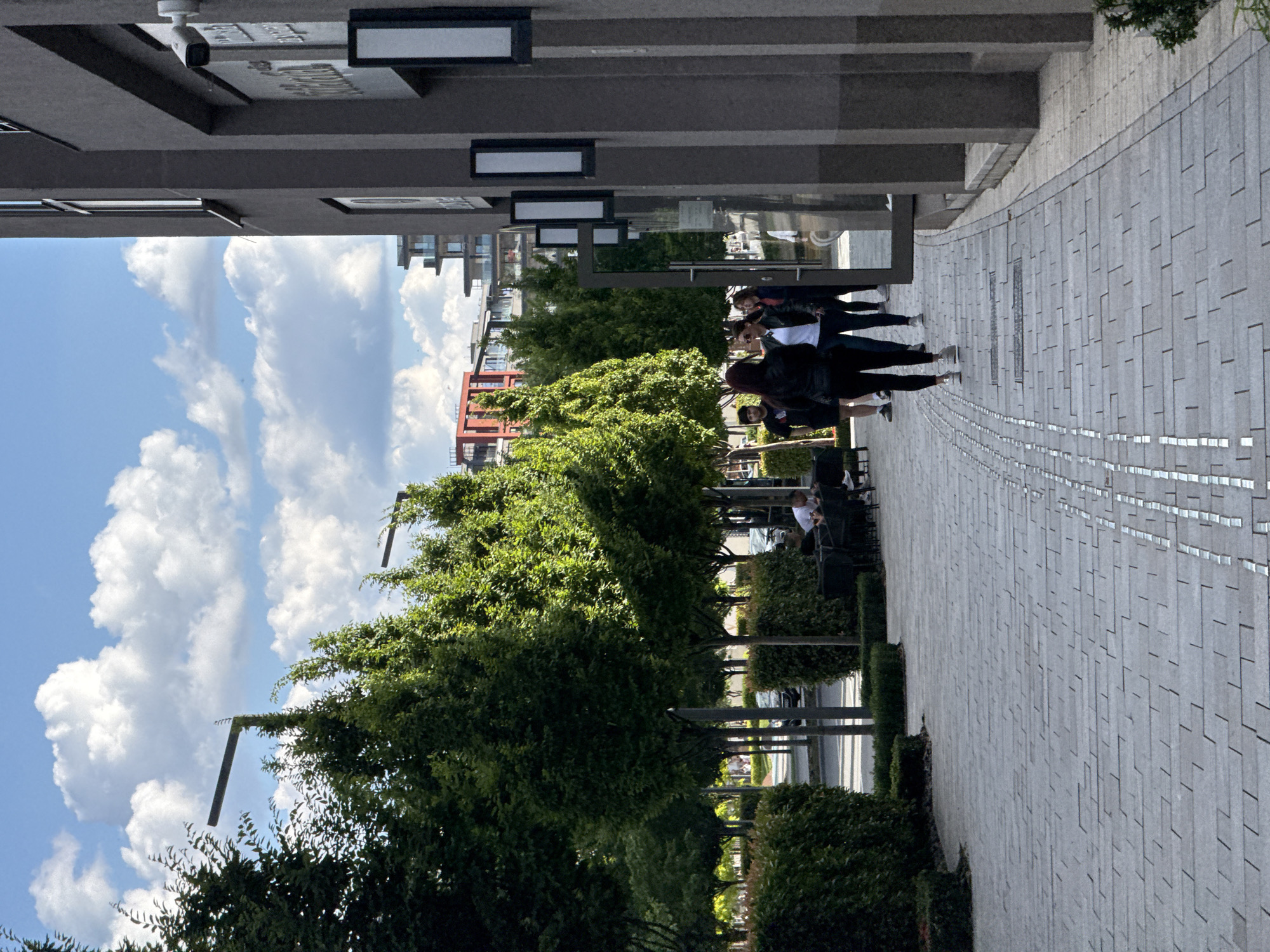 The image depicts an outdoor scene with a paved walkway lined by trees and hedges. On the right side of the walkway is a modern building with a dark facade and rectangular light fixtures. Several people are walking along the pathway, some are sitting at outdoor tables. In the background, there are more buildings, including some modern apartment complexes and a red building. The sky is blue with scattered white clouds. The location is identified as Belgrade, Serbia.
