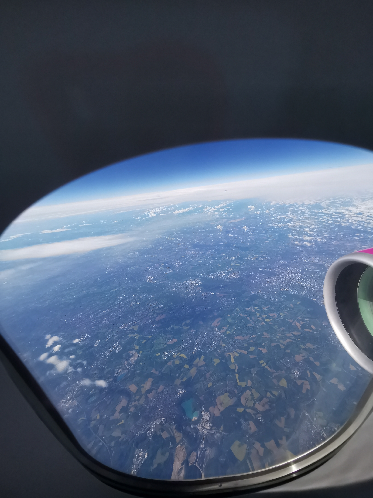 The image is a view from an airplane window, showing the Earth's surface. The horizon is visible as a curved line with a blue sky above and a layer of clouds separating the sky from the land below. The land is a mix of green and brown fields, possibly agricultural land, and there are some visible settlements. Part of the airplane's engine is visible in the lower right corner of the image. The photo was taken during the day.
There is no indication of the location being Varna, Bulgaria, or any information about people, subjects, objects, actions, or interactions.
