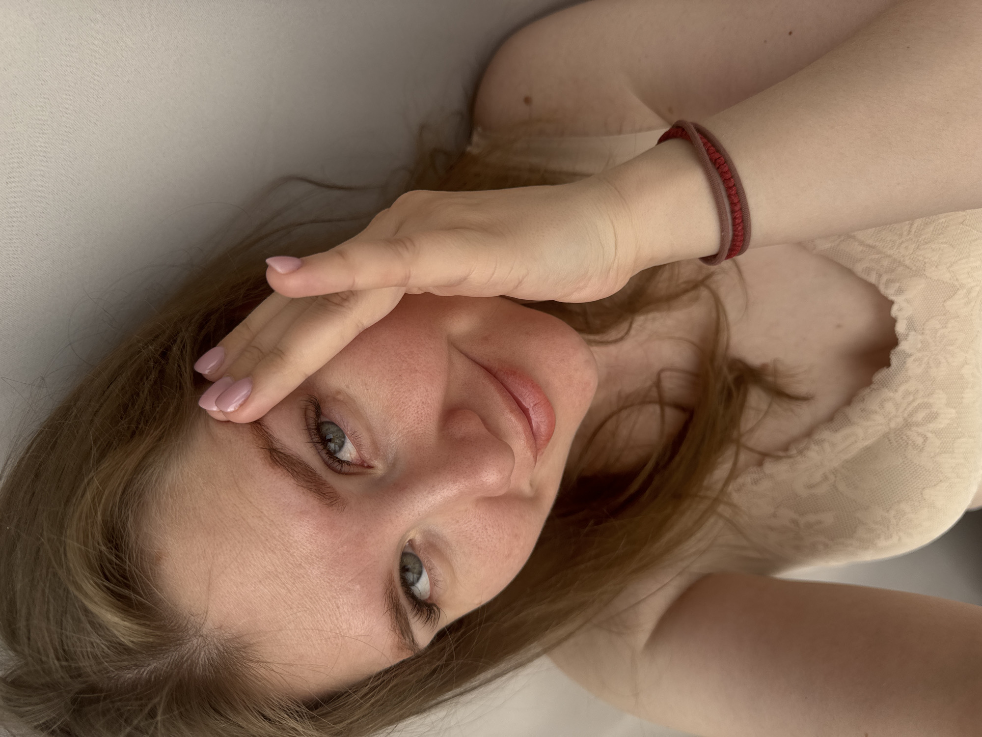 A light-skinned woman with light brown hair and blue-grey eyes is shown in a close-up shot. She is wearing a light beige lace top and a red bracelet on her wrist. She is looking directly at the camera with a slight smile, and her hand is partially covering her forehead. The background is a plain white surface. The location in the city of Gornyi (Dvurechenskoe S/P), Russia is not discernible from the image.