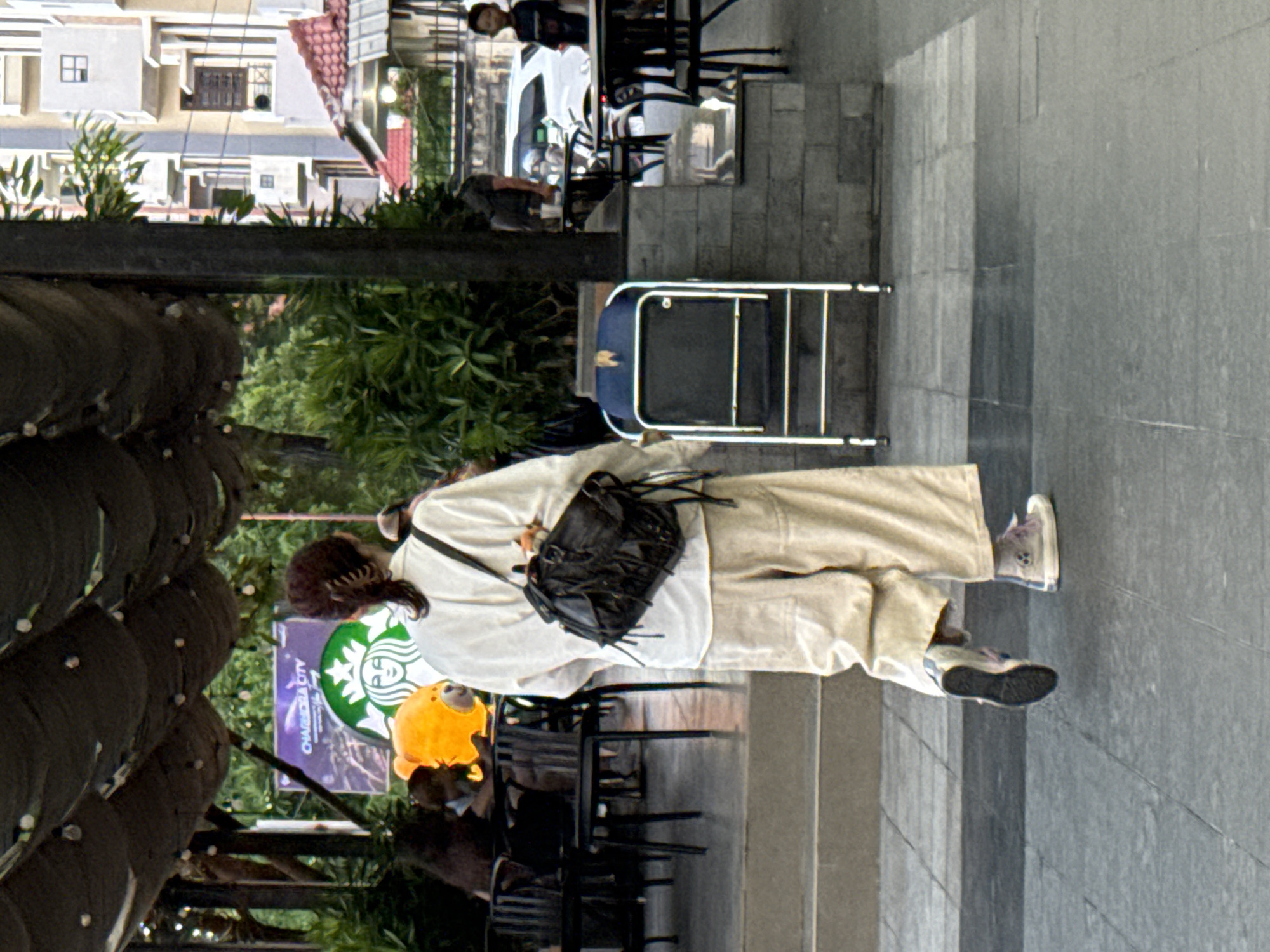 A person walks away from the camera on a paved walkway. The person is wearing loose, cream-colored pants, a white long-sleeved shirt, and white sneakers with purple accents. A dark-colored, fringed shoulder bag is slung across their back. In the background, there are tables and chairs, suggesting an outdoor seating area for a cafe or restaurant. A large Starbucks logo is visible on a sign behind the tables. Further in the background, there are buildings and lush green trees. The overall scene appears to be an outdoor public space in a city.