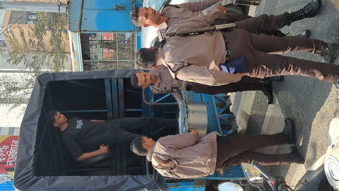 A scene shows a flatbed truck carrying a young man wearing a black shirt, lying down inside. The truck appears to be stopped on the street with four uniformed police officers standing around it. Three of the police officers are grouped near the front of the truck, while one stands slightly apart. They are on a pedestrian crossing. A building is in the background.