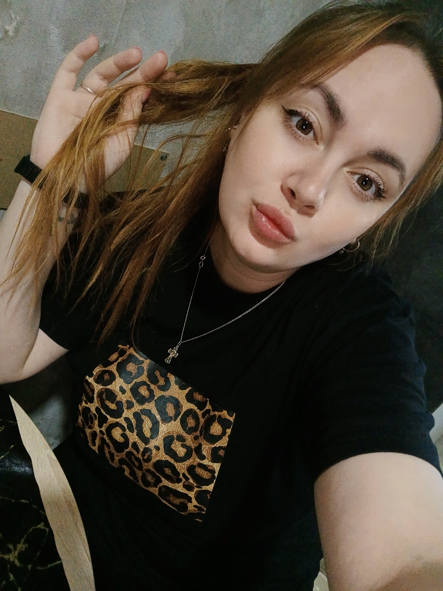 The image shows a selfie of a young woman with reddish-brown hair. She is holding a strand of her hair in her left hand, displaying a silver ring on her finger and a black wristwatch on her wrist. She has brown eyes with eyeliner and her lips are pursed slightly. She wears a silver necklace with a cross pendant. She is wearing a black shirt with a leopard print pocket square. The background is nondescript and light-colored. There is no visible information to confirm the location in Chaikovsky, Russia.