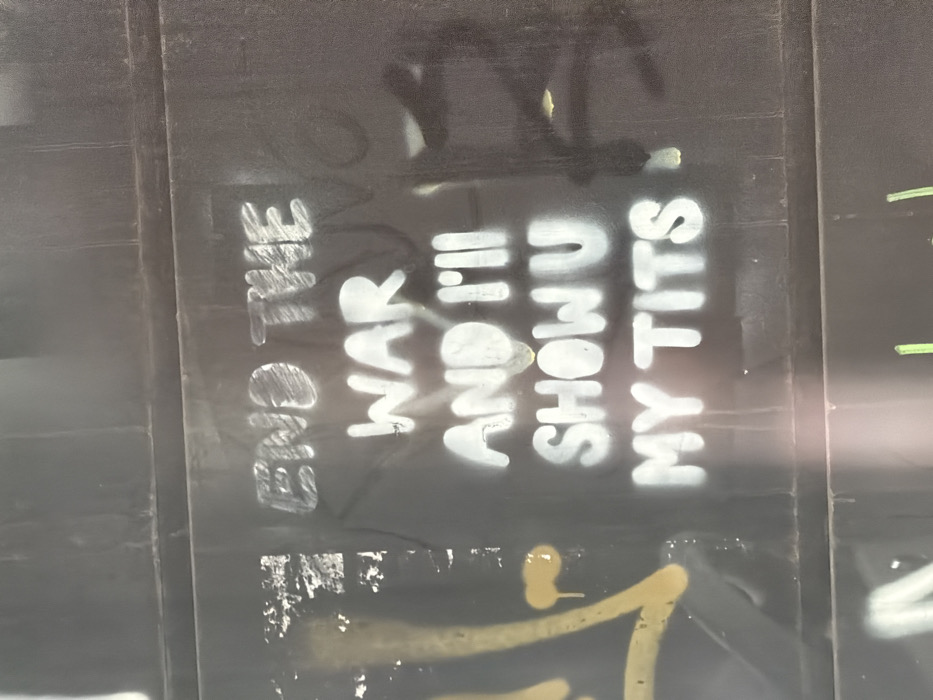 The image displays a dark, multi-paneled wall or barrier located in Tel-Aviv, Israel, covered in graffiti. The dominant feature is a message spray-painted in white capital letters, utilizing a stencil effect, which reads: "END THE WAR AND I'LL SHOW U MY TITS." This text is centrally positioned on a dark gray or black background that shows wear and subtle surface variations. To the left of the white text, fragments of yellow spray paint are visible, including drips and parts of abstract shapes. Faint, darker markings or older graffiti layers are discernible beneath and around the prominent white message. A horizontal seam or structural division spans the width of the wall in the lower half of the frame. A small section of green spray paint is visible at the bottom edge. No people are present in the scene.