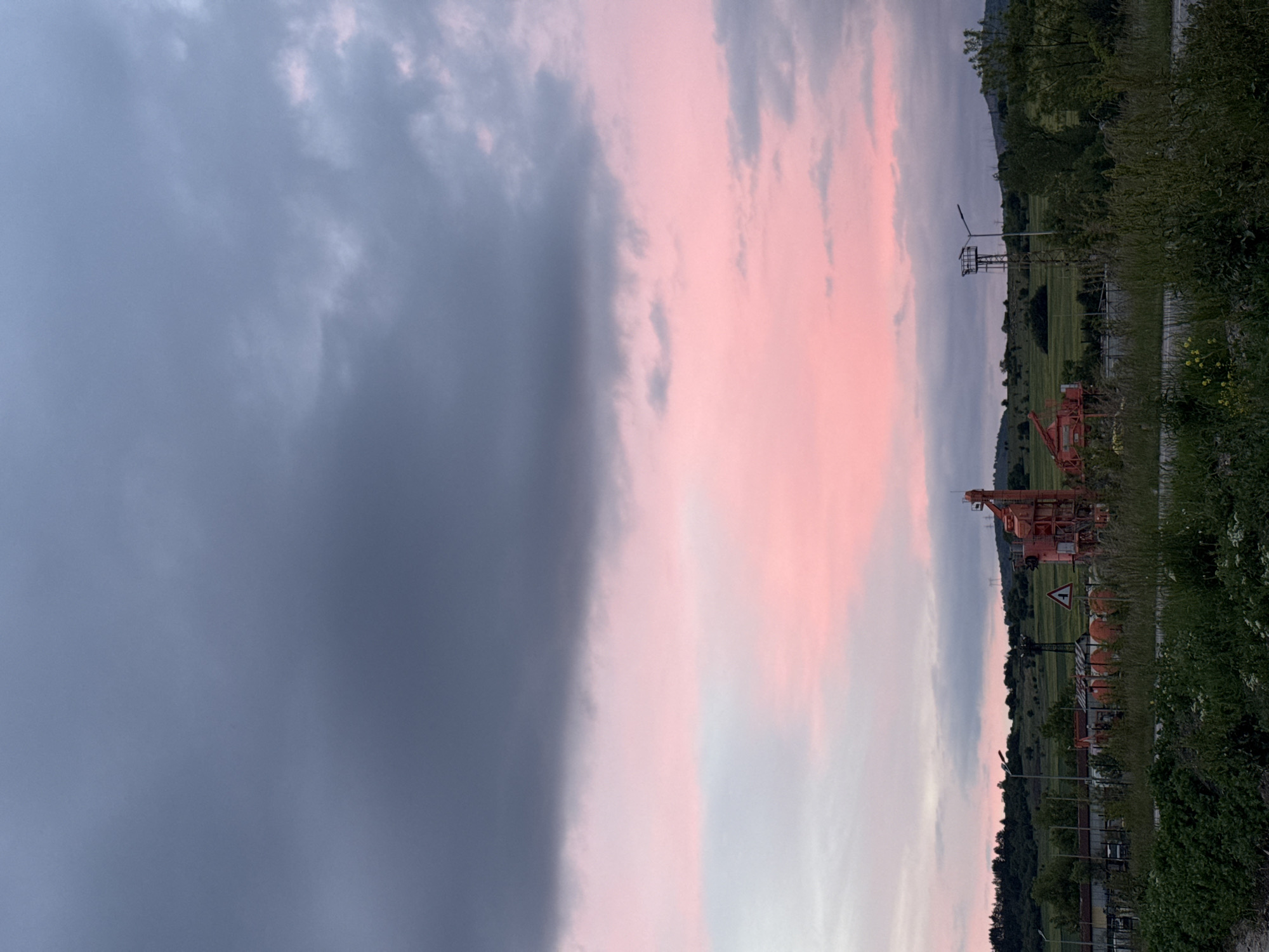 The image shows a landscape with a contrasting sky. On the left, a large, dark gray cloud formation dominates the view. To the right of this cloud, the sky transitions into hues of pink and light blue, suggesting a sunset or sunrise. The ground is covered with greenery, including grass and trees. In the middle distance, there are some structures, including a metal tower and what appears to be construction equipment. The photograph is taken from ground level. The location is Unknown, Unknown.