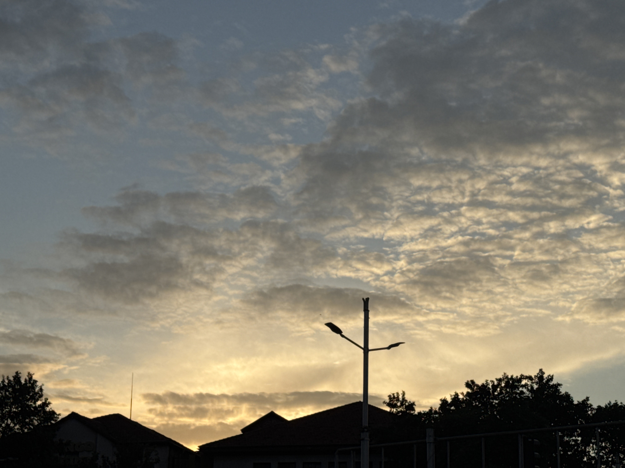The image captures a sky filled with scattered clouds during what appears to be sunrise or sunset, with the lower portion of the frame dominated by silhouetted rooftops and trees. A prominent street lamp stands near the center of the image, its dark form stark against the illuminated sky. The overall mood is peaceful and serene, with the muted colors of the sky and the dark silhouettes creating a dramatic contrast. The location is identified as Plovdiv, Bulgaria, though no specific landmarks within the city are discernible in this general cityscape view. The absence of any visible people or specific actions suggests a moment of stillness.