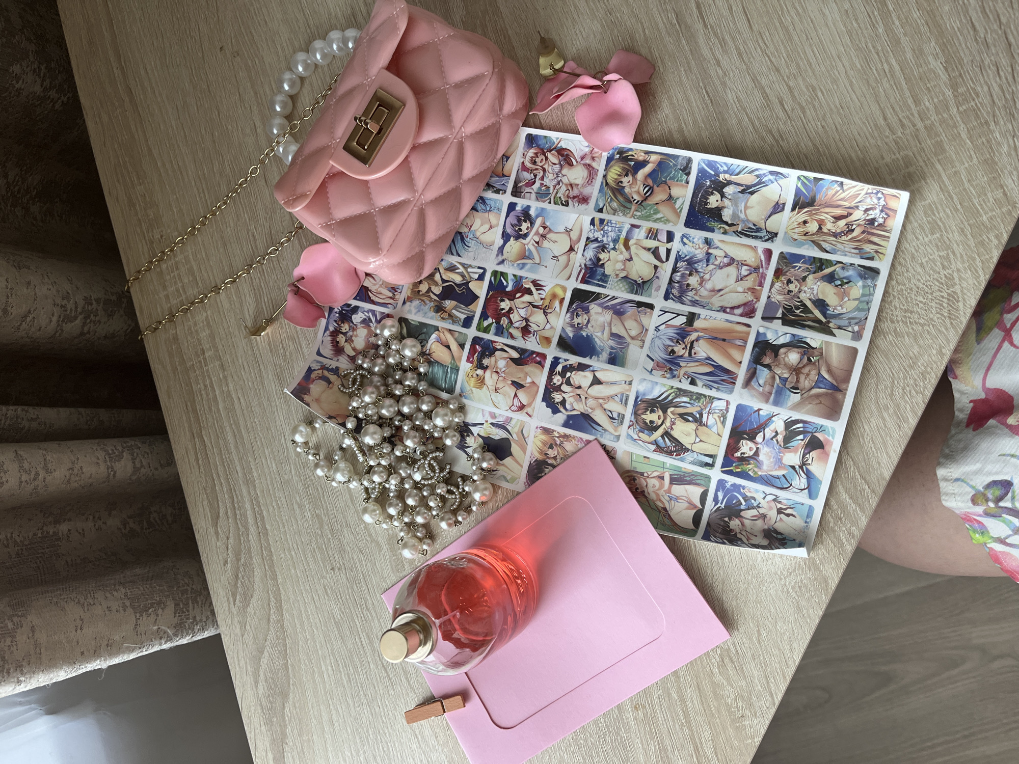The image shows a flat lay of feminine items on a wooden surface. The objects include a pink envelope with a wooden clothespin, a glass perfume bottle with pink liquid, a strand of pearls, a small quilted pink purse with a gold chain, and a sheet of stickers featuring anime-style illustrations of women in bikinis. The background consists of a neutral-colored curtain and a wooden table. The scene is well-lit, suggesting it was taken indoors during the daytime. There is no visible indication of the location being in Kazan, Russia, or any other specific geographical place.