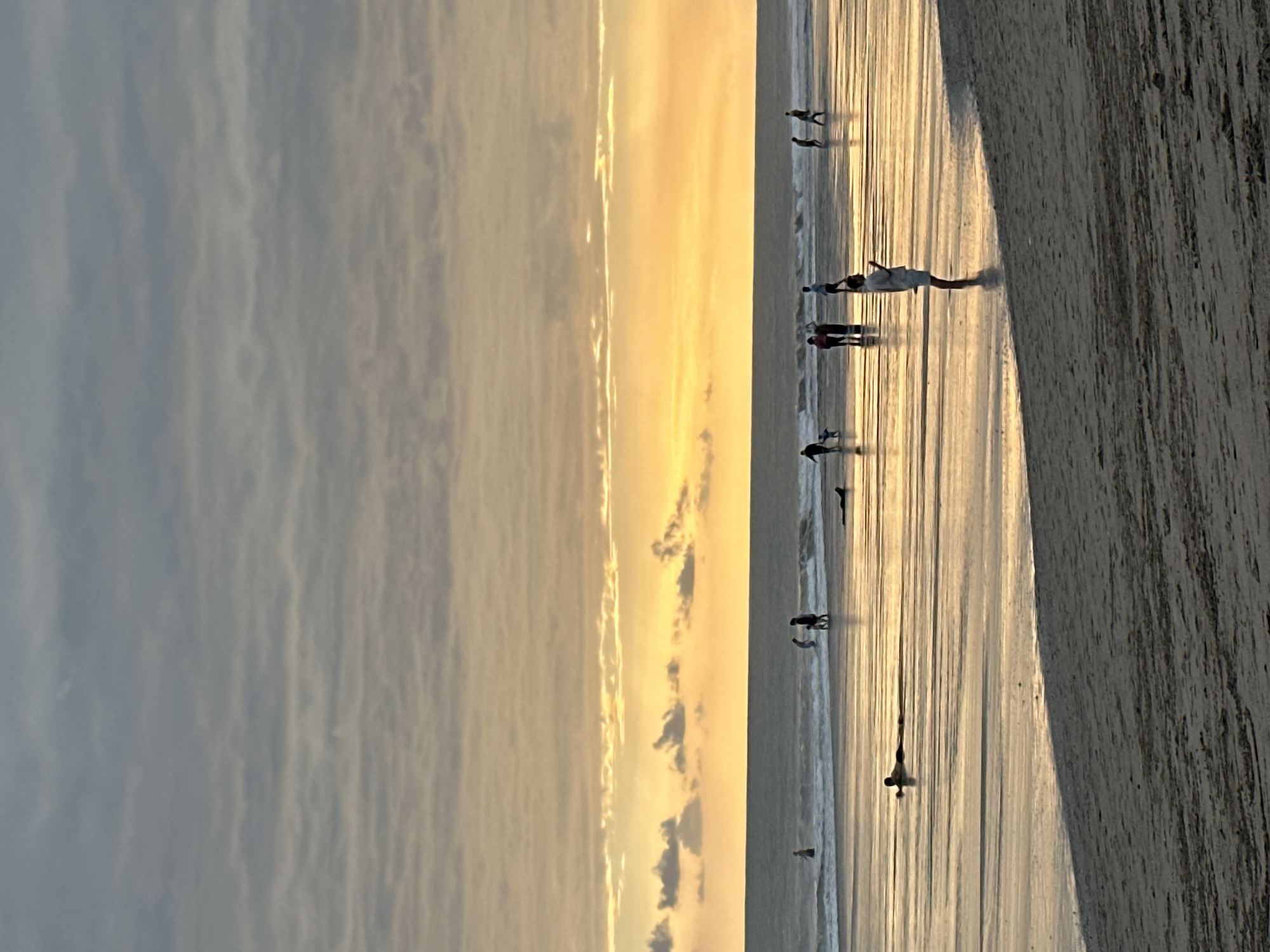 This is a photograph of a beach scene during sunset in North Kuta, Indonesia. The sky is filled with bands of clouds in varying shades of grey, blue, and orange, with a bright orange glow near the horizon. The ocean is visible, with small waves rolling onto the shore. The wet sand reflects the colors of the sky and the people on the beach.
There are several people visible on the beach and in the shallow water. In the foreground, a person is standing on the wet sand, facing away from the camera, wearing light-colored clothing. Further out in the water, a group of people are standing and walking. To the left, a person is lying on their stomach in the shallow water, facing the camera. In the distance, more figures can be seen walking along the shoreline and in the water. The overall impression is a serene evening at the beach.