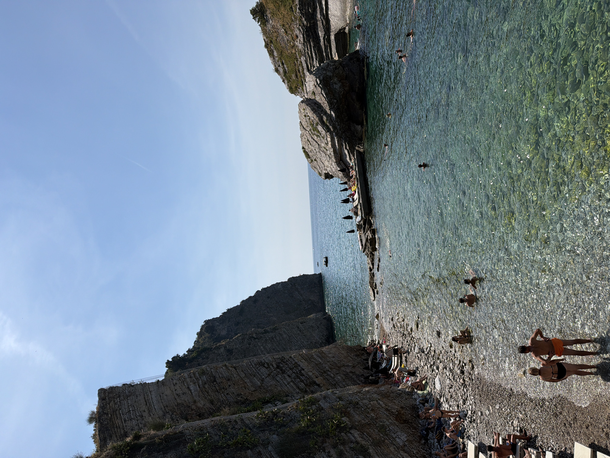 The image depicts a rocky cove with a pebble beach, situated near a cliff face in Budva, Montenegro. The clear, turquoise water teems with swimmers of various ages. On the shore, a mix of sunbathers and beachgoers are visible. Some are seated on towels or lounge chairs, while others stand or walk along the water's edge. A couple stands in the shallow water in the foreground, facing away from the camera. Further into the cove, a group of people relax on sun loungers under umbrellas on a small concrete platform. The scene is bathed in bright sunlight under a clear blue sky.