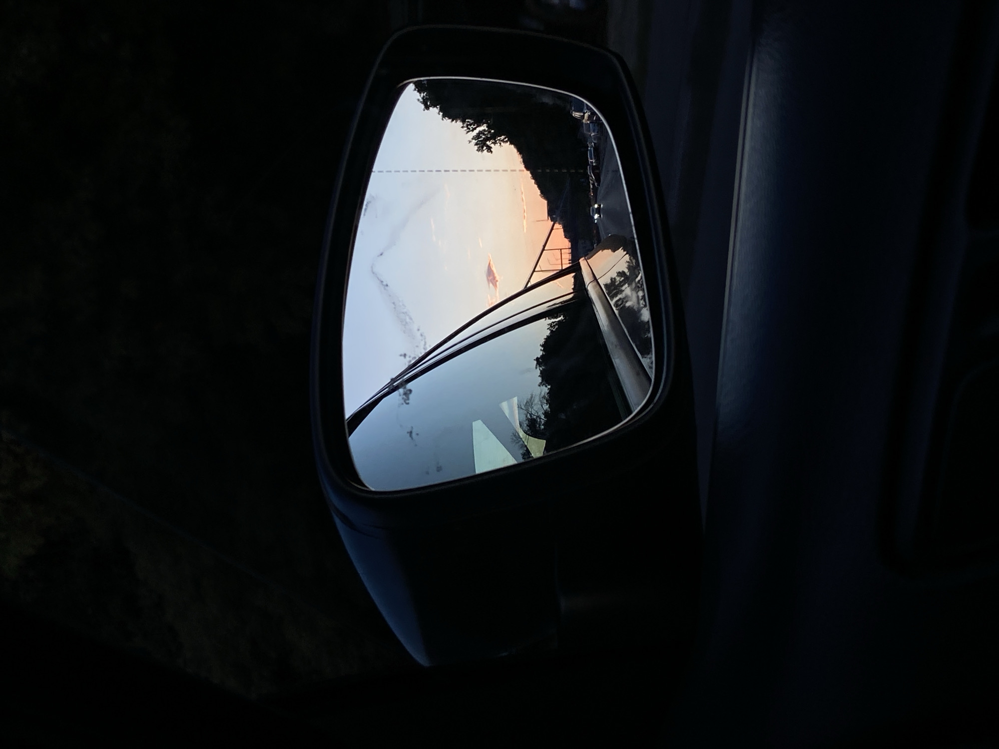 A close-up shot focuses on the right-side wing mirror of a car. The mirror reflects a scenic view of a sunset on a highway. The sky is a gradient of orange, pink, and blue, with a few wispy clouds. A bridge spans across the highway in the distance. The highway is populated with a line of cars, their headlights illuminated. Trees and a hillside are visible on the right side of the reflection, and the car's side mirror housing is dark and looms in the foreground on the left. The overall ambiance is one of twilight travel.