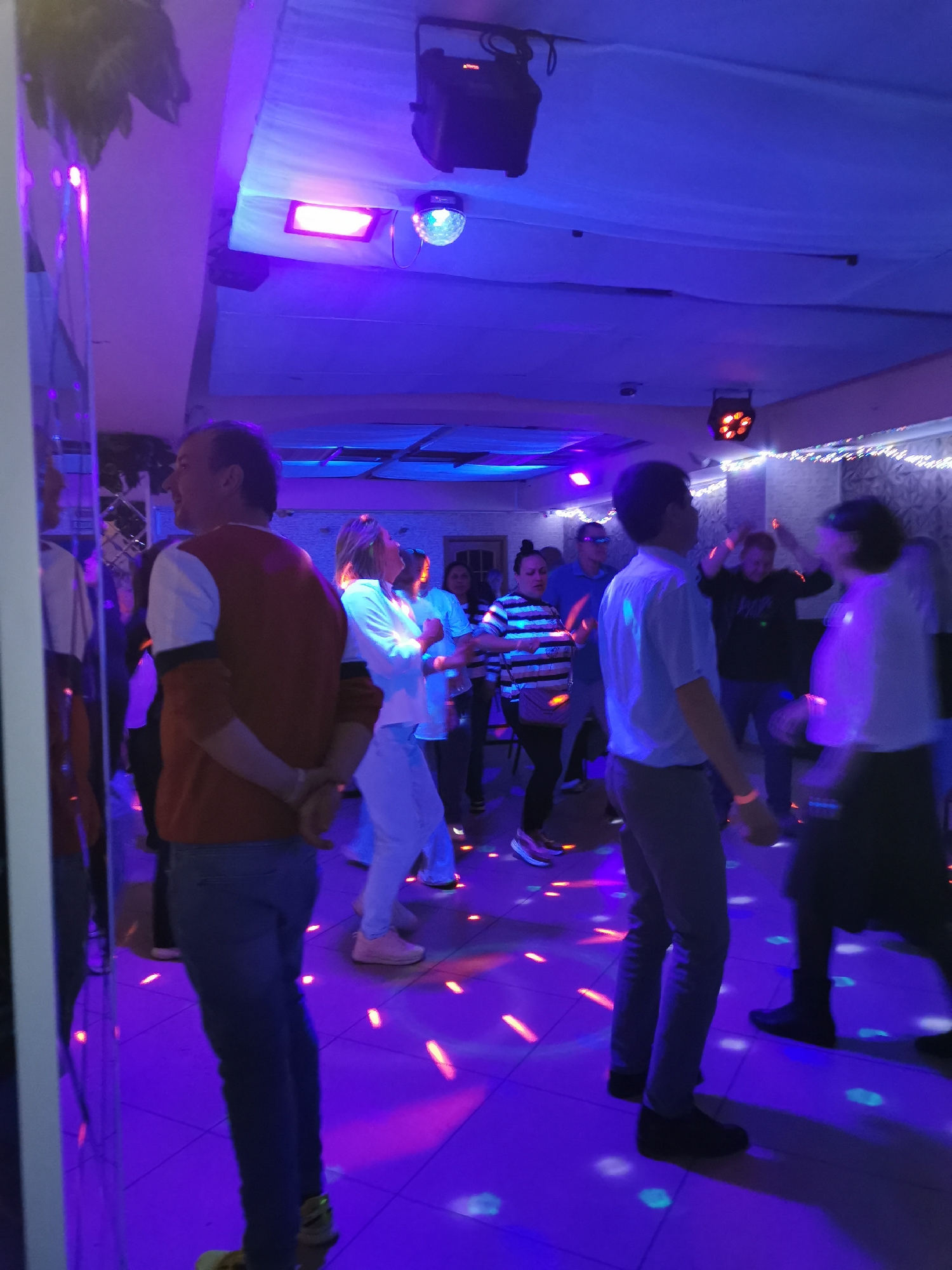 This media file captures an indoor party or club scene, illuminated by vibrant purple and blue disco lighting. Numerous people, both men and women of varying ages, are visible throughout the space, engaged in dancing or socializing. The atmosphere appears lively and energetic, suggested by the motion blur on several individuals, indicating movement.
In the foreground to the left, a man with short brown hair, wearing a rust-colored and white long-sleeved shirt and blue jeans, stands with his hands clasped behind his back, looking towards the right side of the room. A reflective, possibly mirrored, surface is present along the left edge of the frame, showing partial reflections of people.
The ceiling is draped with light-colored fabric, from which several lighting fixtures hang, including a rectangular red light panel and a rotating disco ball projecting colorful patterns onto the floor and attendees. Another multi-lens light fixture, emitting an orange glow, is visible further back on the ceiling. The floor is covered in square tiles, onto which red, blue, and white light patterns are projected.
Further into the room, other party-goers are visible: a woman in a white long-sleeved top and light-colored pants; a man in a light blue short-sleeved shirt; a woman in a black and white striped shirt; and several other figures in the background, some with their arms raised in dance. The walls are decorated with textured or patterned wallpaper. No discernable text is visible in the image. The lighting suggests the event is taking place at night or in a dimly lit indoor environment.
