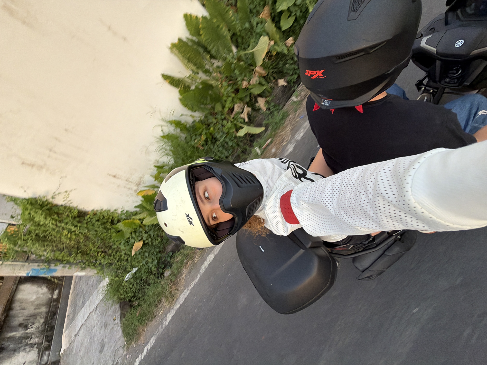 In North Kuta, Indonesia, two individuals are captured from a first-person perspective, riding on a motorcycle. The person in the foreground, wearing a white mesh long-sleeved shirt with red accents on the wrist and a light-colored helmet with a black face mask, extends their arm. The person in the background, wearing a black t-shirt and a black helmet with red accents, is visible to the right. They are on a road bordered by a concrete wall with vegetation and a dirt path. The image appears to have been taken during daylight, with the sun casting a warm glow.