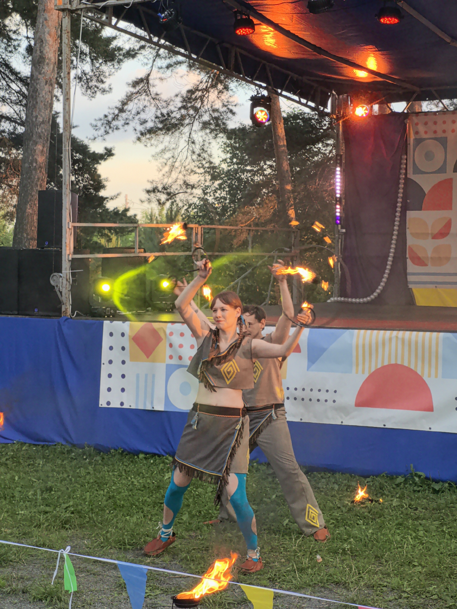 This image shows two performers on a stage with fire props in the city of Gorniy, Russia. The stage is set up outdoors, likely for a festival or event, with trees and a twilight sky in the background. The performers are dressed in costumes that appear to be inspired by tribal or folk attire, featuring earthy tones, fringe details, and geometric patterns. One performer, a woman in the foreground, is actively spinning fire, creating a bright halo of flames around her. She wears a brown vest-like top, a skirt, and teal leggings with cutouts. The second performer, a man to her right, is also holding a prop that emits fire. He wears a similar outfit. Both performers appear focused on their act. In front of the stage, there is a blue banner with a colorful geometric design. The ground is covered in green grass, with some fire effects on the ground as well. The lighting on the stage is a mix of theatrical lights and natural twilight, casting a warm glow.