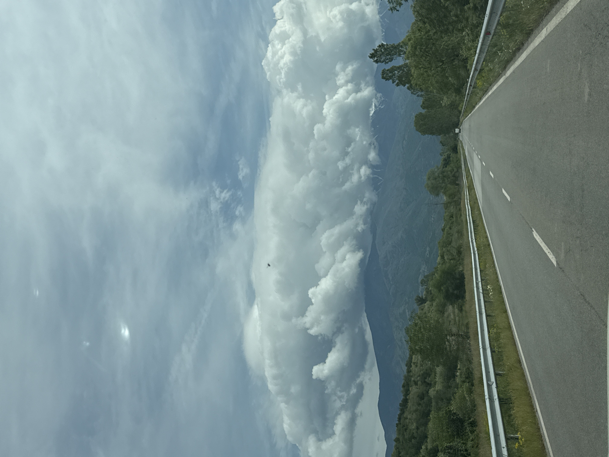 A straight asphalt road with a white dividing line stretches into the distance. A silver guardrail lines the right side of the road. To the right of the road is green vegetation. To the left of the road are distant mountain ranges and a large, vertical cumulus cloud. The sky is pale blue. The road may be near Kocherinovo, Bulgaria.